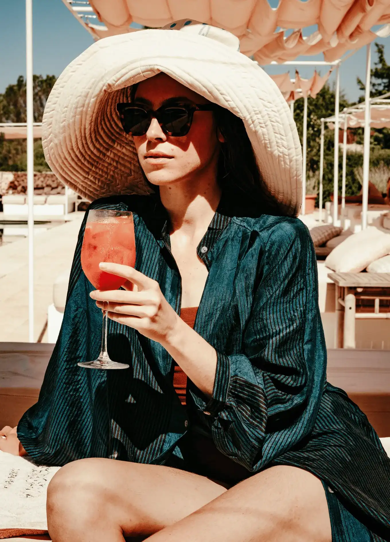 Donna che indossa un grande cappello e occhiali da sole che tiene un cocktail rosso all'aperto in una zona lounge soleggiata.