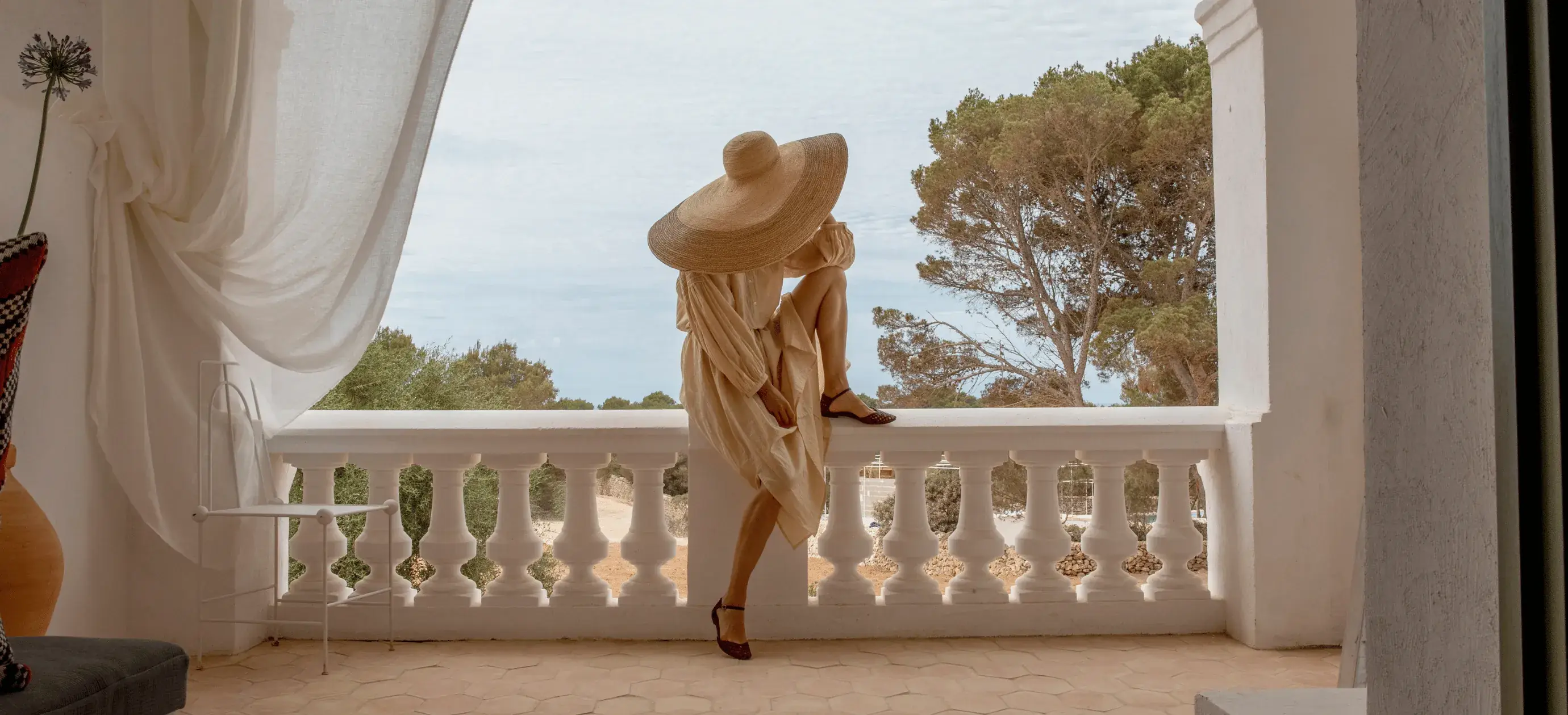 Donna in abito beige e grande cappello seduta su una ringhiera bianca del balcone con alberi sullo sfondo.