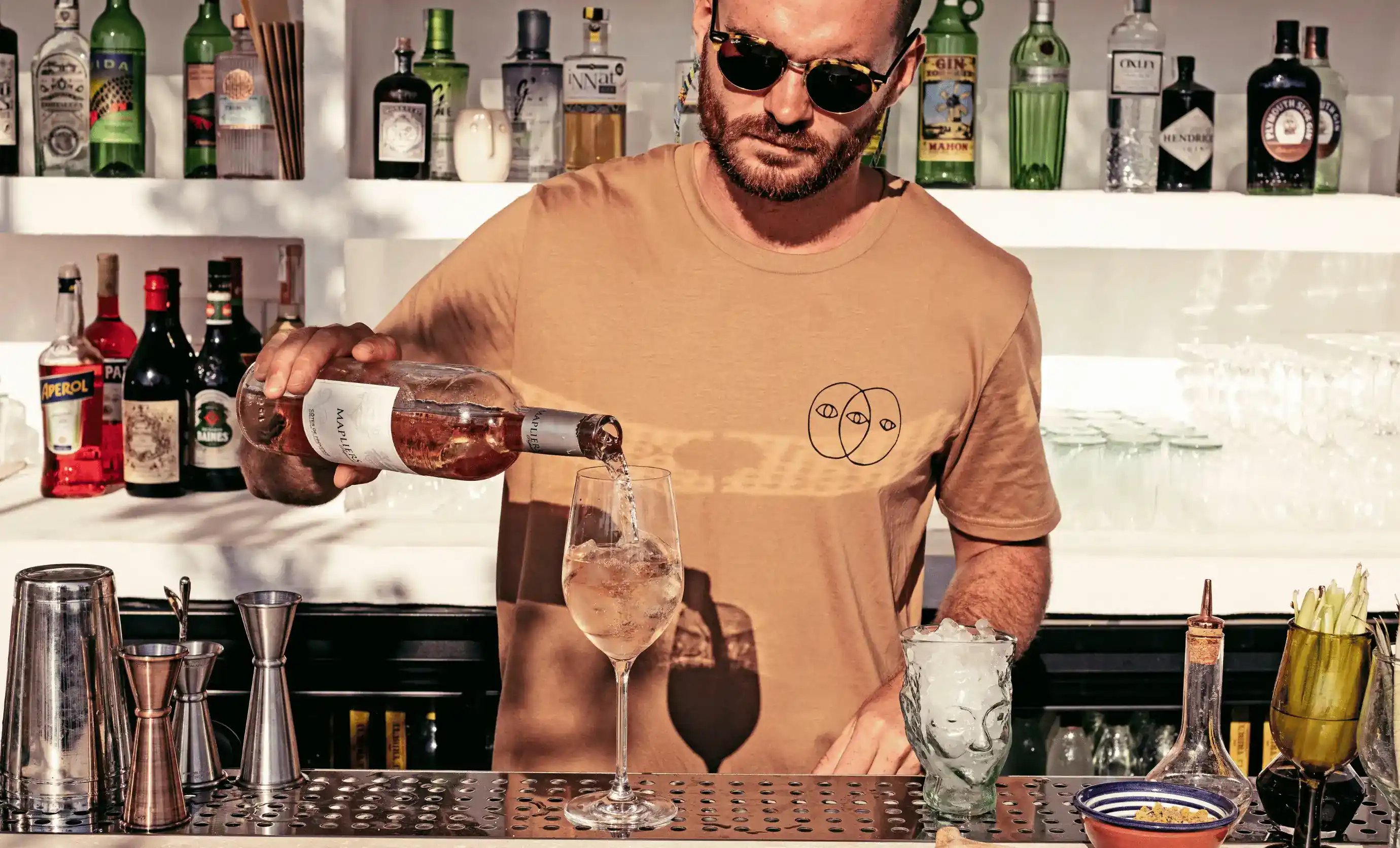 Barista con occhiali da sole che versa vino rosato in un bicchiere dietro un bar con bottiglie.
