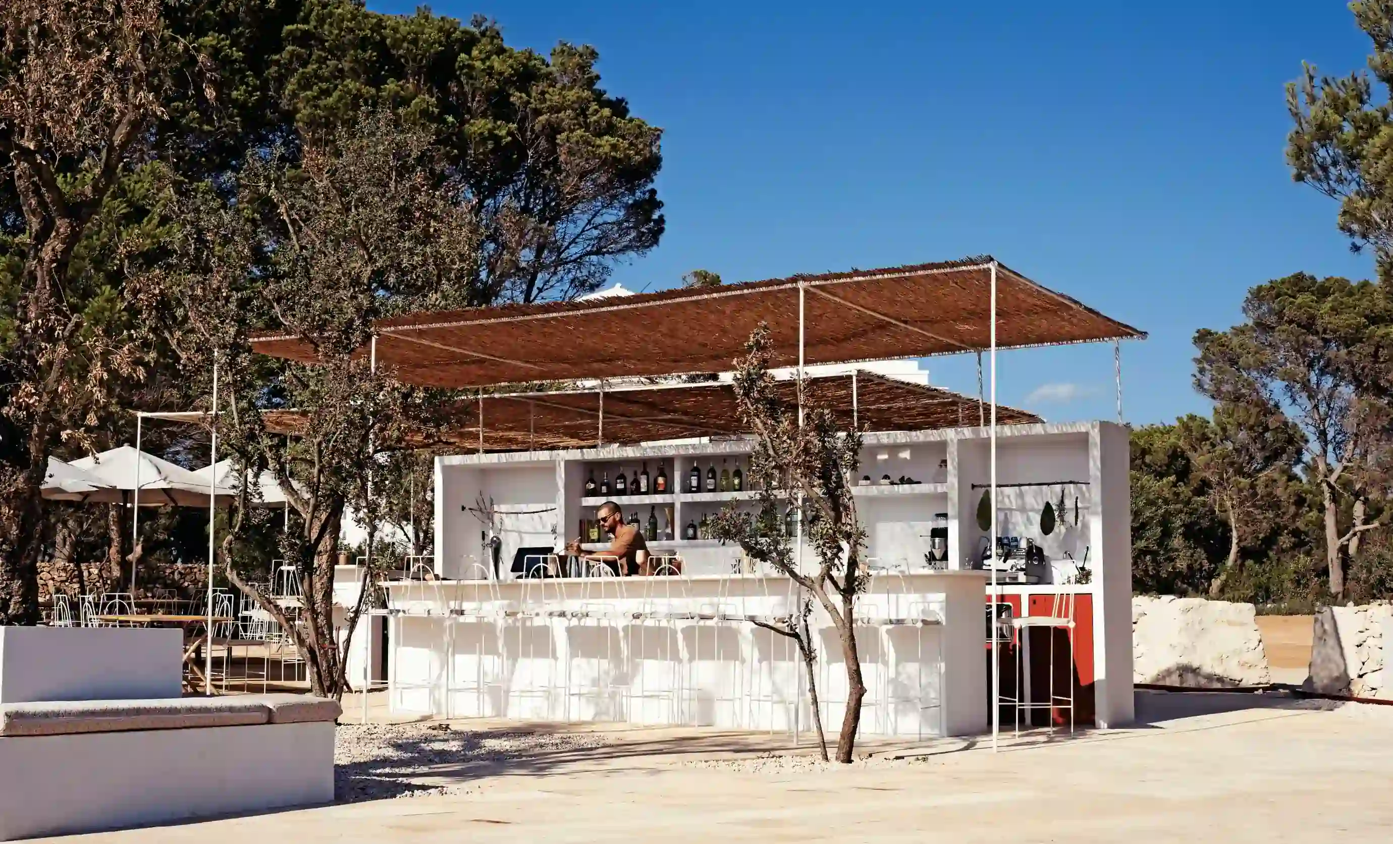 Bar all'aperto con sgabelli bianchi, tettoia, barista e alberi circostanti sotto un cielo blu chiaro.