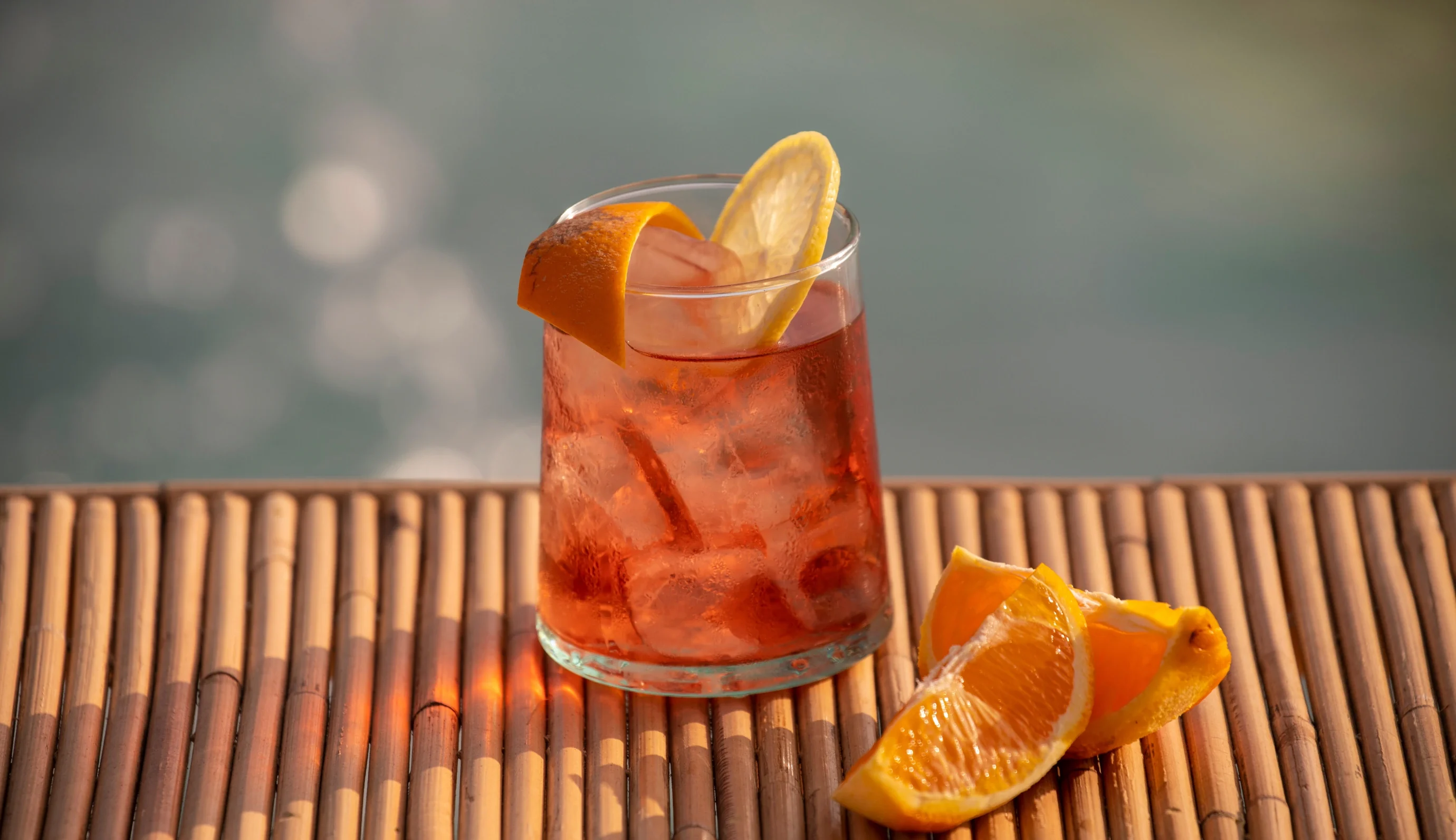 Bicchiere con cocktail rosso ghiacciato guarnito con fetta di limone e spicchi d'arancia su stuoia di bambù.