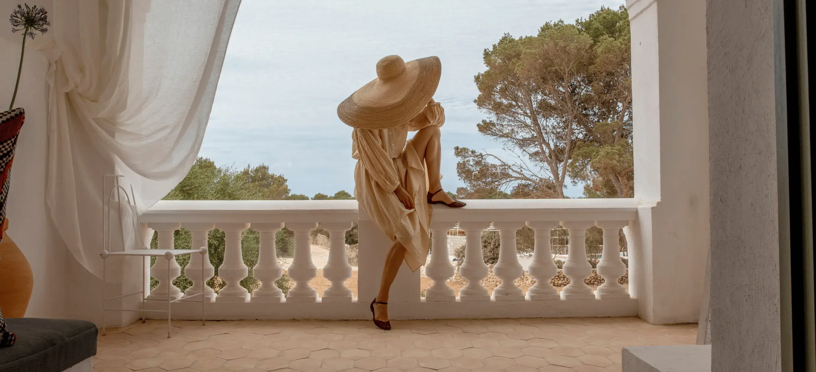 Donna in abito beige e cappello largo seduta su una ringhiera bianca del balcone con vista sugli alberi.
