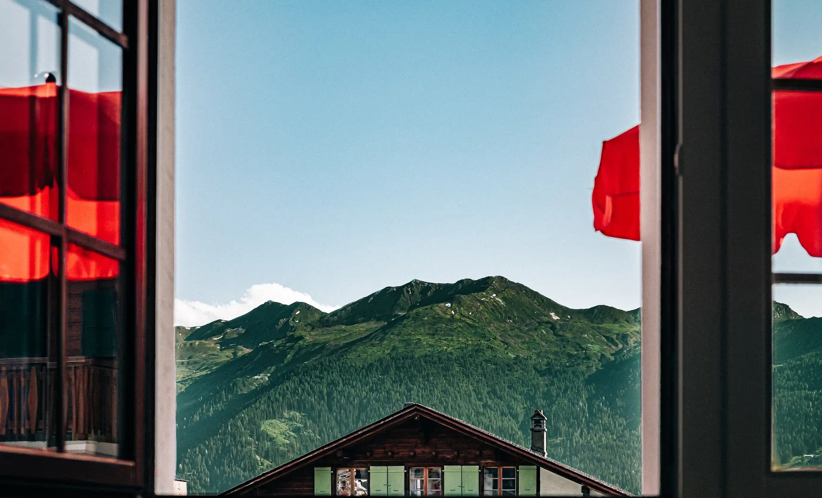 Vista sulle montagne con foresta verde e tetto dello chalet visto attraverso una finestra con tende rosse.