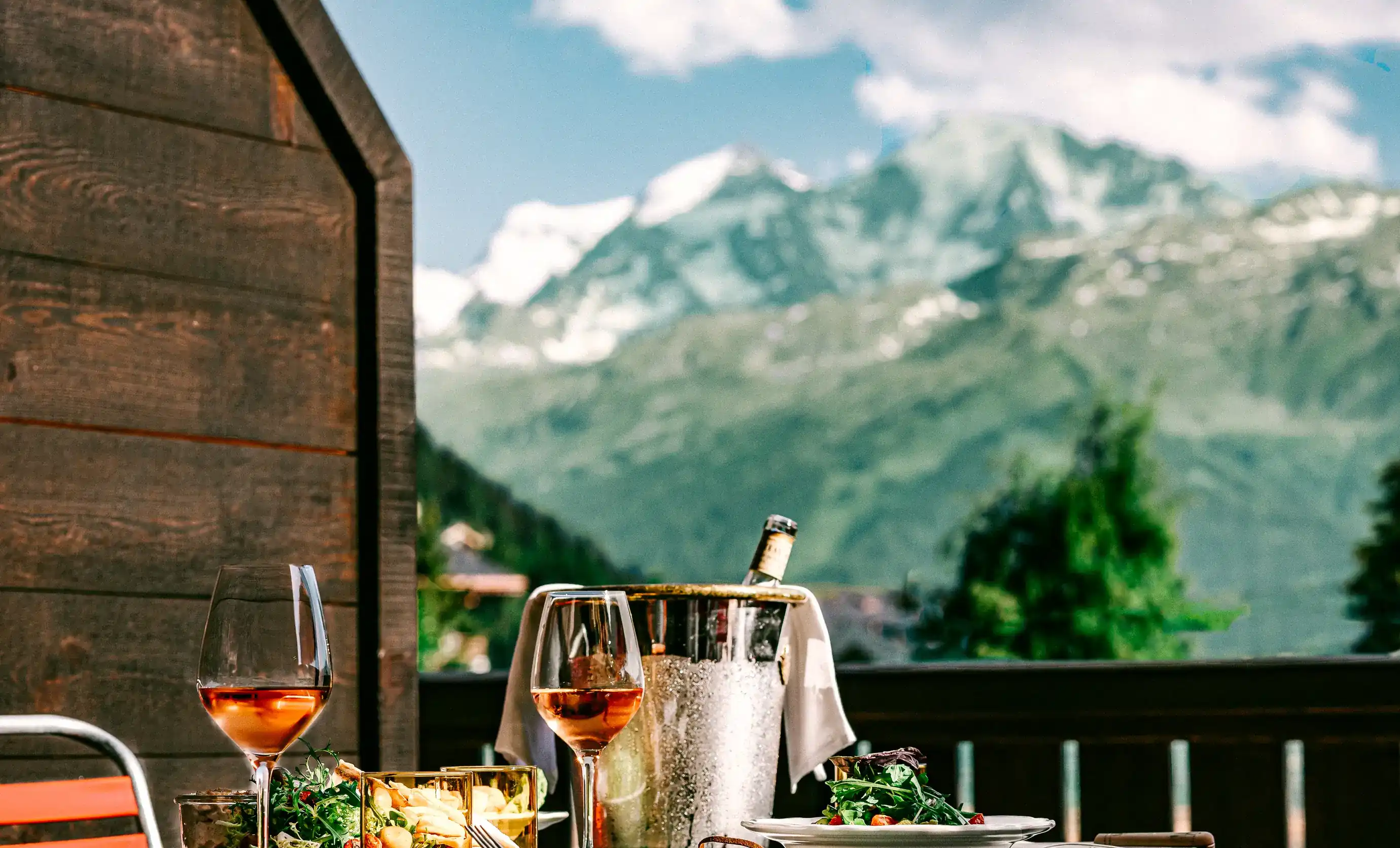 Due bicchieri di vino rosato, piatti di insalata e bottiglia di vino in un secchiello per il ghiaccio su una terrazza in legno con vista sulle montagne.