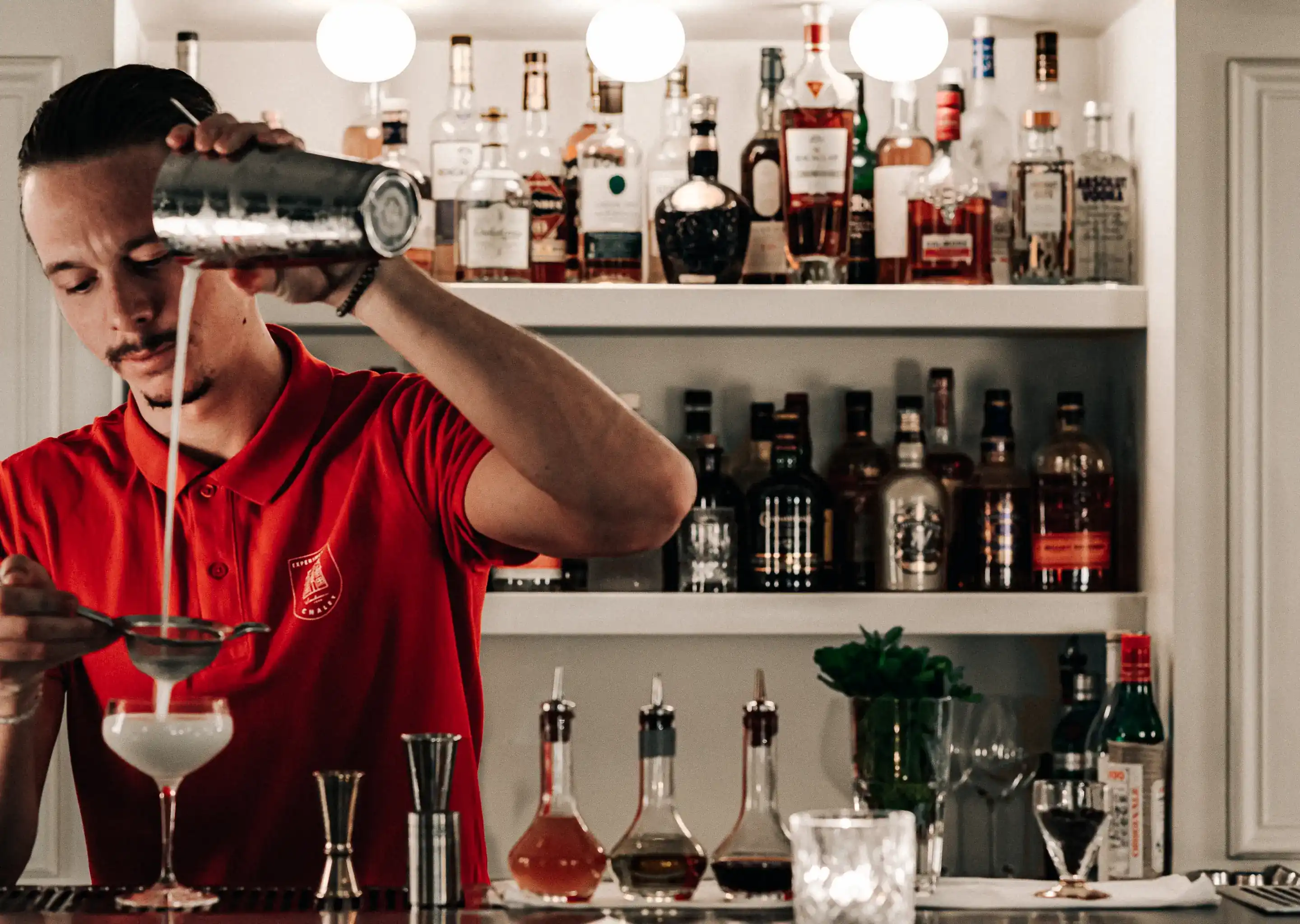Il barista con una camicia rossa versa un cocktail attraverso un colino in un bicchiere da coupé dietro il bancone.