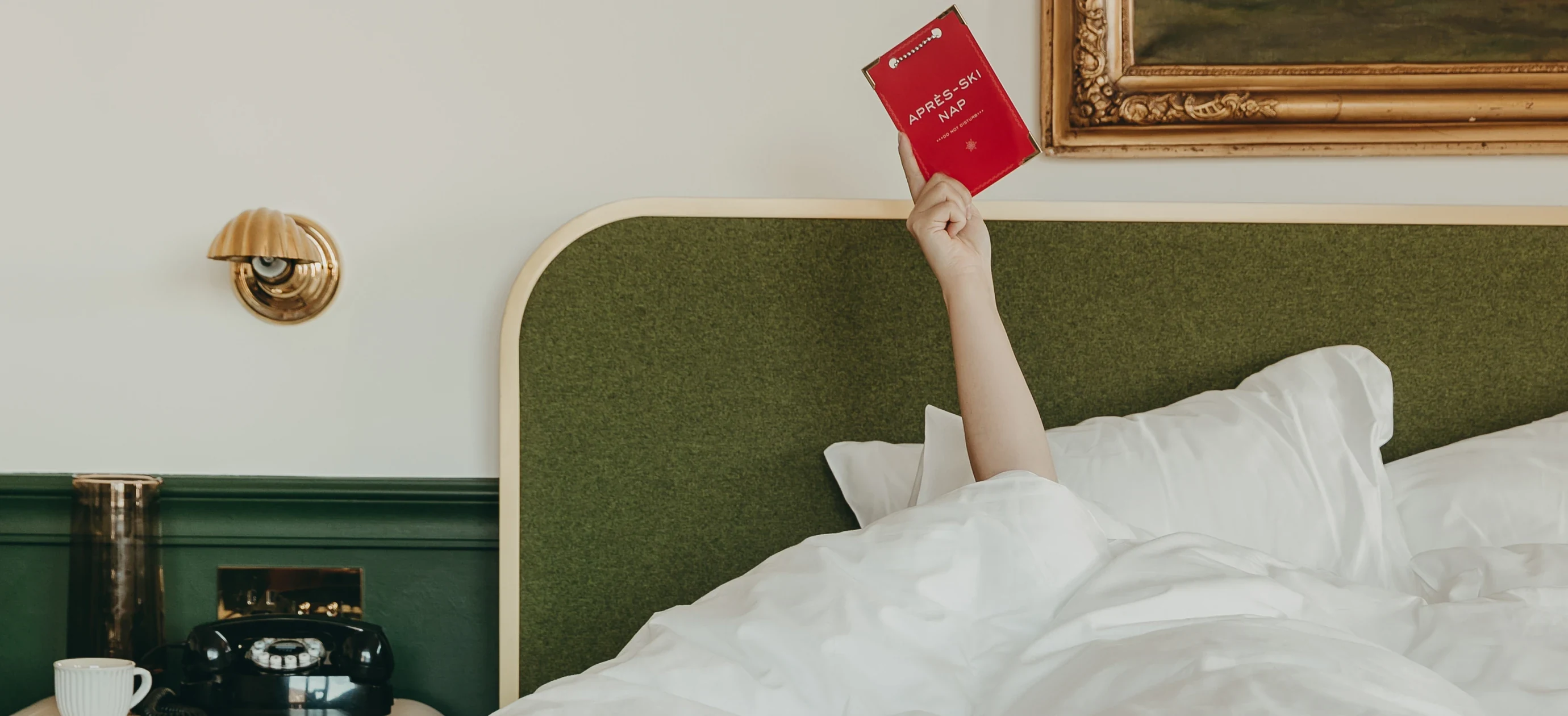 Persona a letto sotto coperte bianche con in mano un cartello rosso con la scritta «APRES-SKI NAP» sopra una testiera verde.