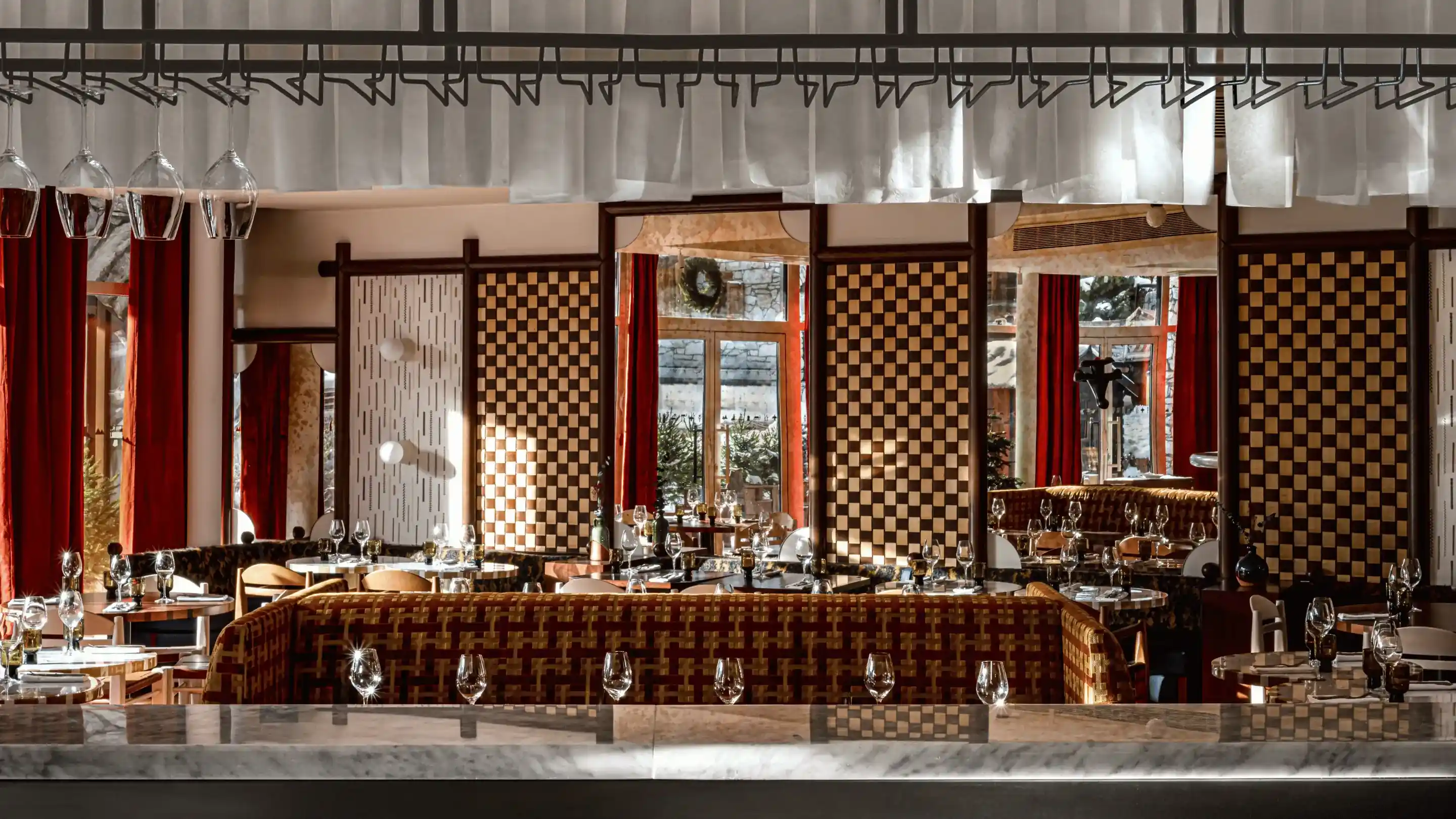 Interior do restaurante com painéis de parede quadriculados, cortinas vermelhas, assentos variados e taças de vinho nas mesas.