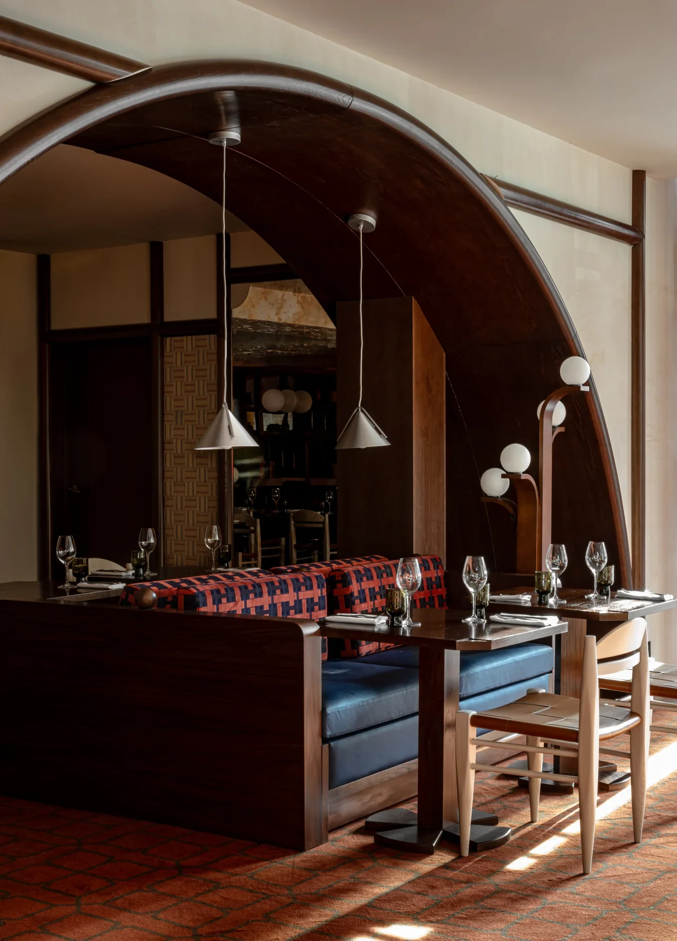 Assentos de restaurante com mesas de madeira, banco estampado, cadeiras, luzes pendentes e luminárias de globo sob um arco curvo de madeira.