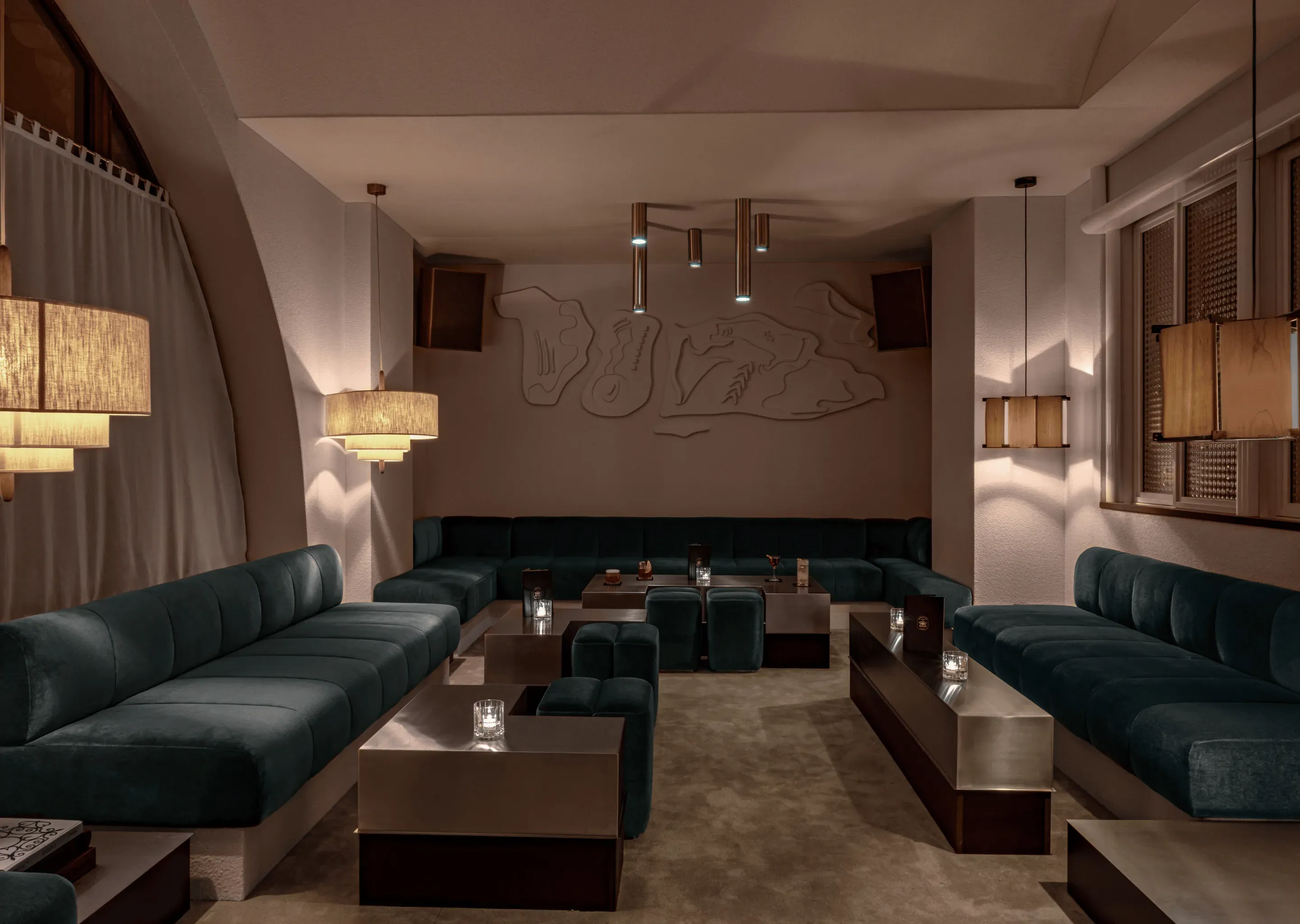 Lounge mal iluminado com sofás de veludo azul-petróleo, luminárias suspensas modernas e arte abstrata na parede em um ambiente aconchegante.