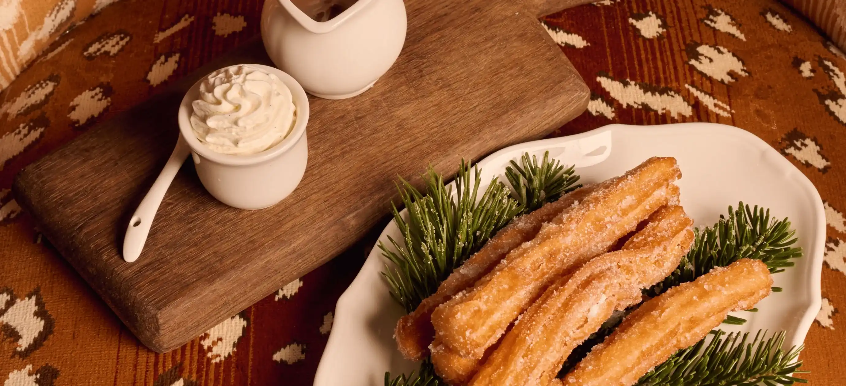 Prato de churros cobertos de açúcar em galhos de pinheiro com chantilly e molho em tábua de madeira.