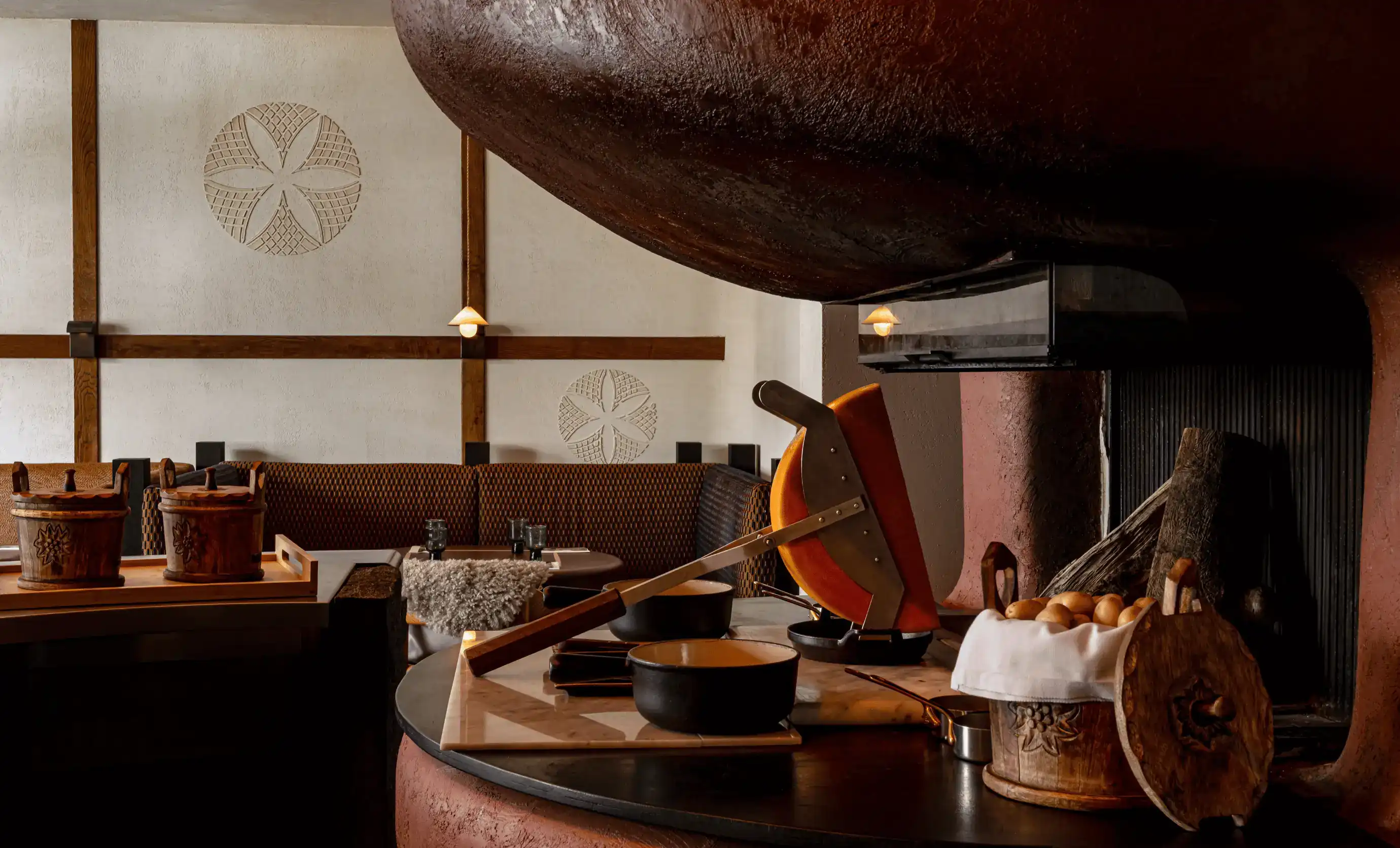 Área de jantar rústica com uma grande roda de queijo derretido, panelas pretas e recipientes de madeira em um balcão.