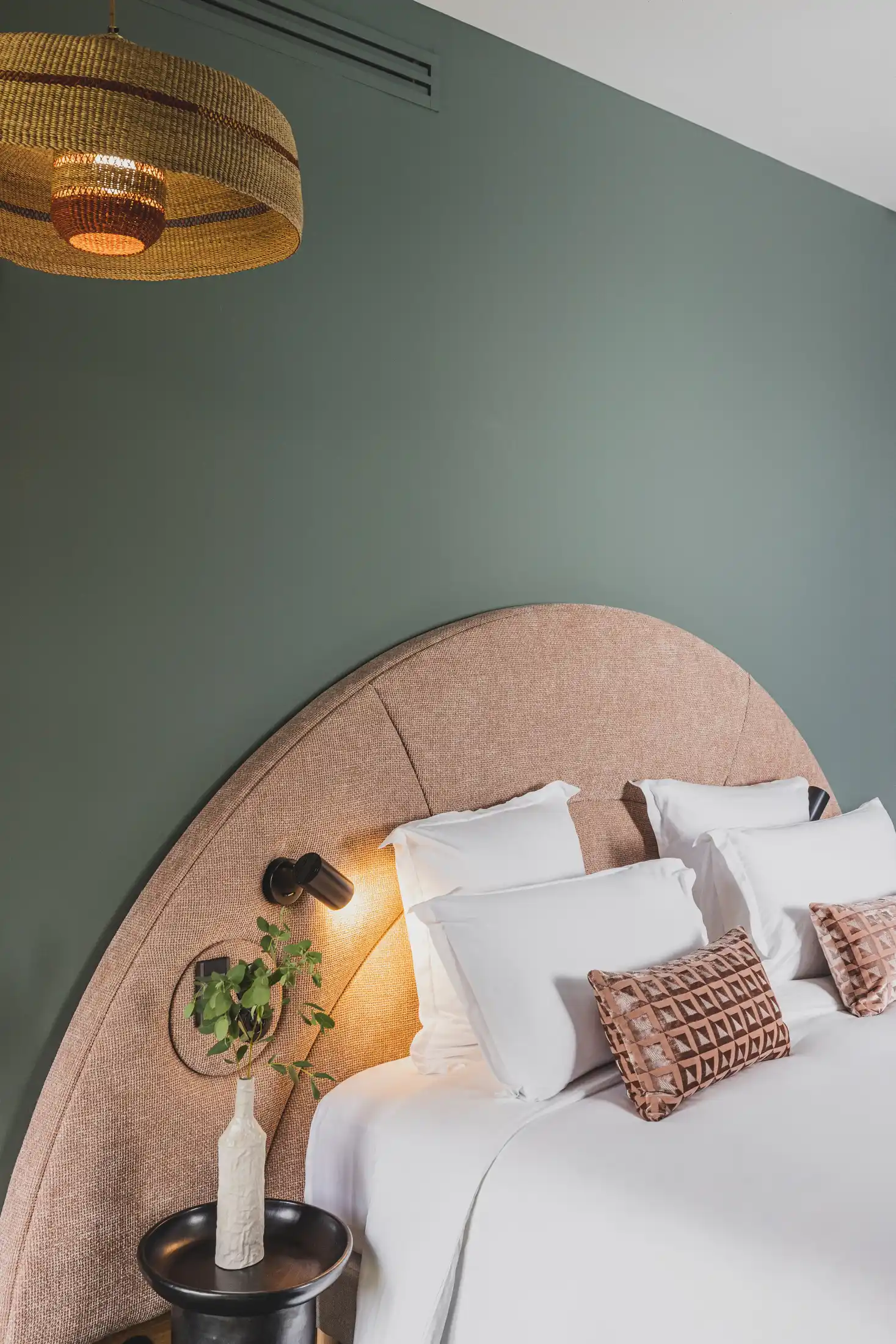 Cama con cabecera grande de tela curvada, ropa de cama blanca, almohadas decorativas y una mesa auxiliar con un jarrón.