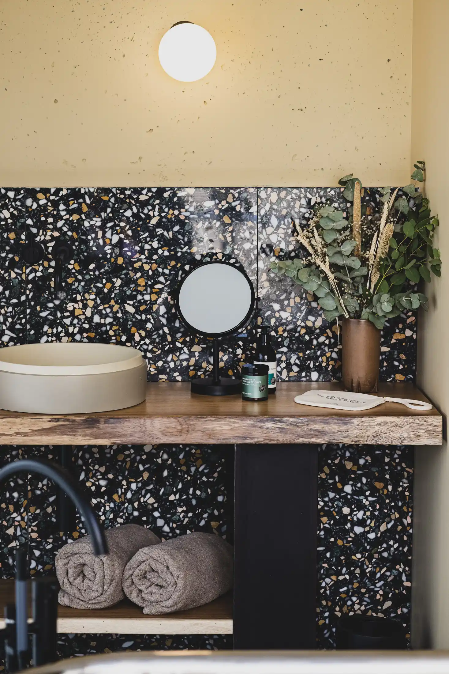 Moderna encimera de baño con lavabo redondo, espejo redondo, artículos de tocador, flores secas y toallas enrolladas en la parte inferior.