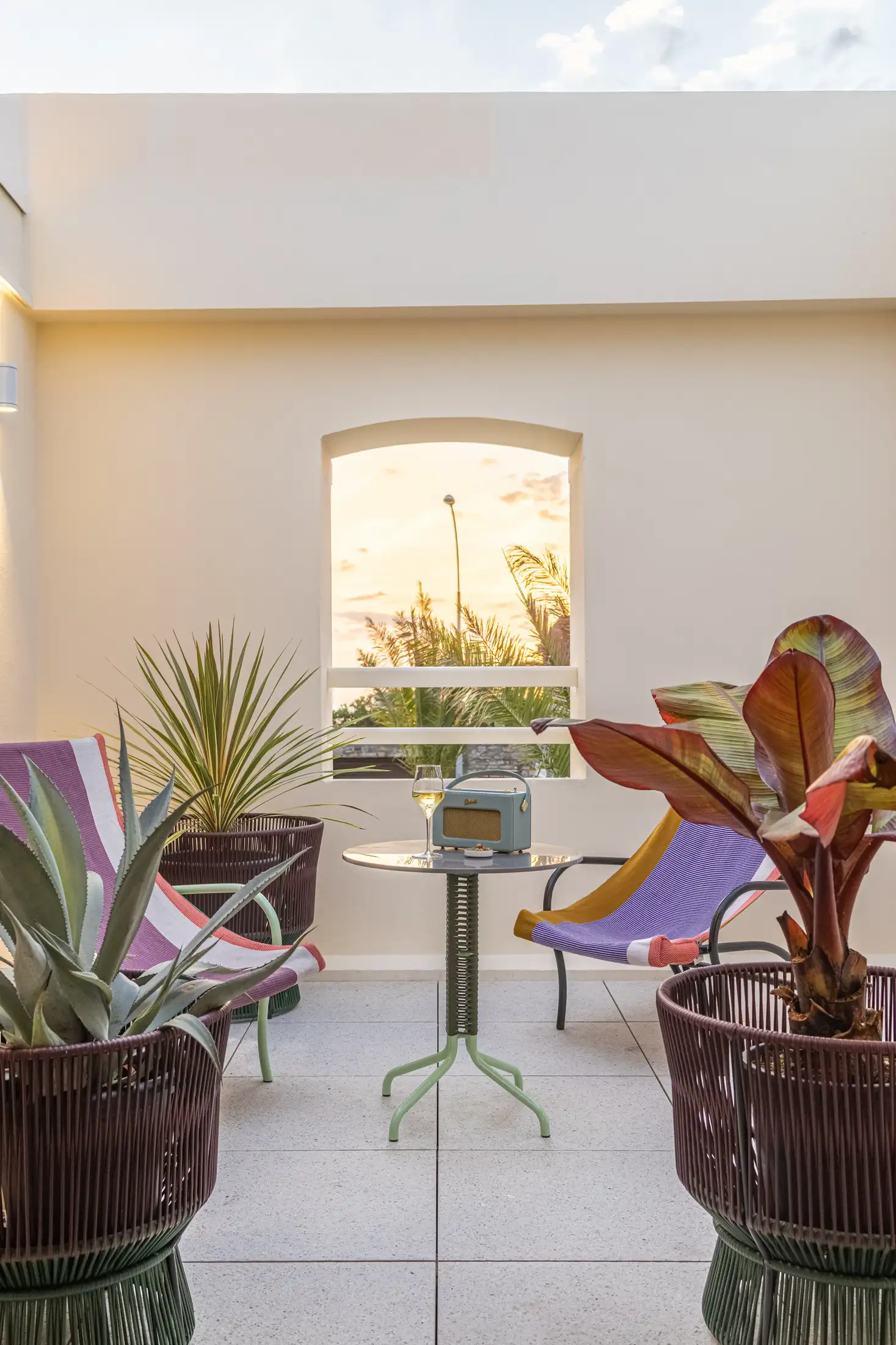 Patio iluminado por el sol con dos sillas de colores, una pequeña mesa verde, plantas y una radio con una copa de vino.