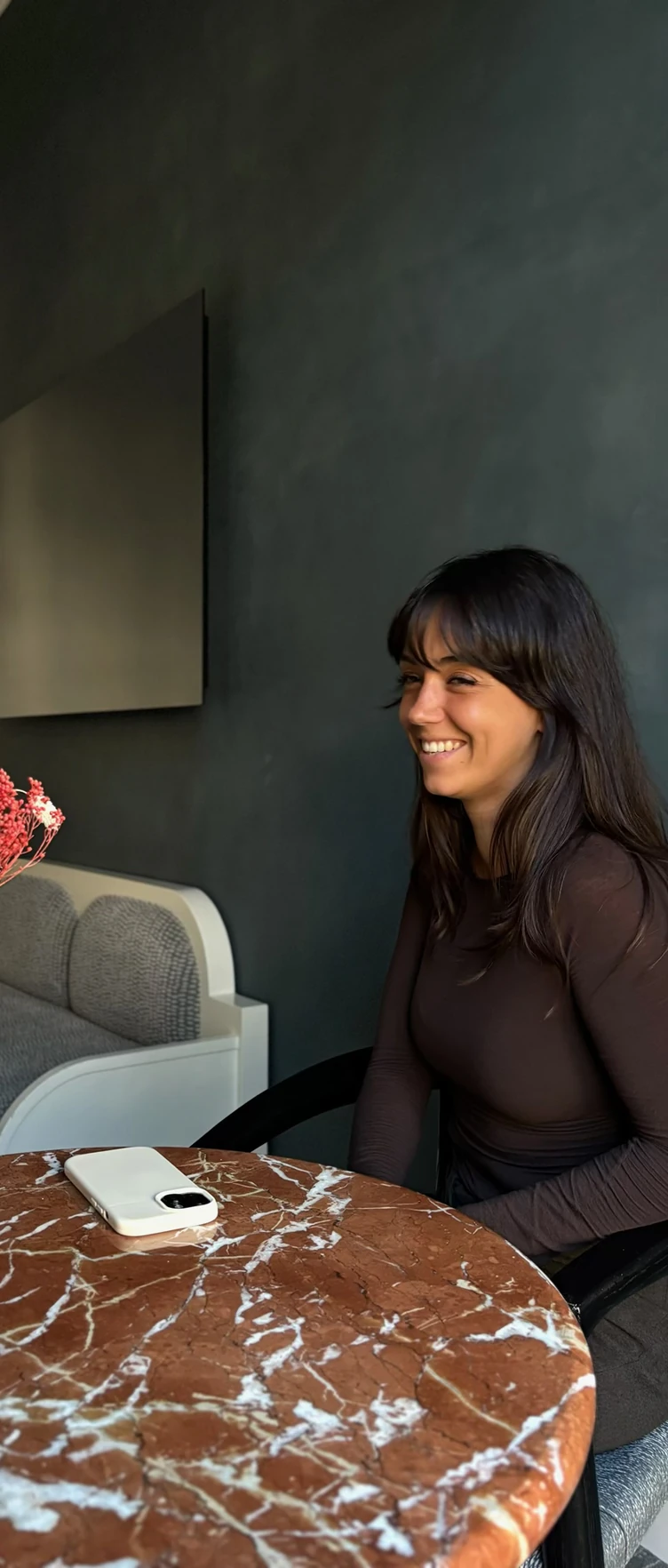 Smiling woman with dark hair sitting at a round brown marble table with a phone on it.