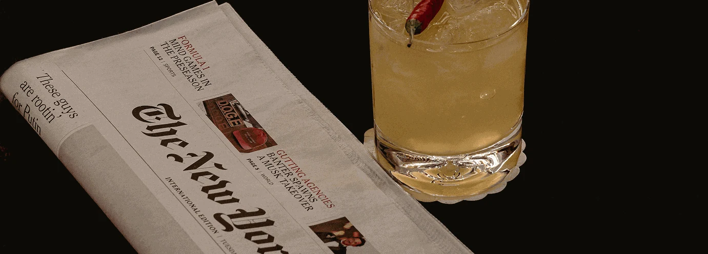 Cocktail giallo con guarnizione di peperoncino rosso accanto a un giornale piegato del New York Times su una superficie scura.
