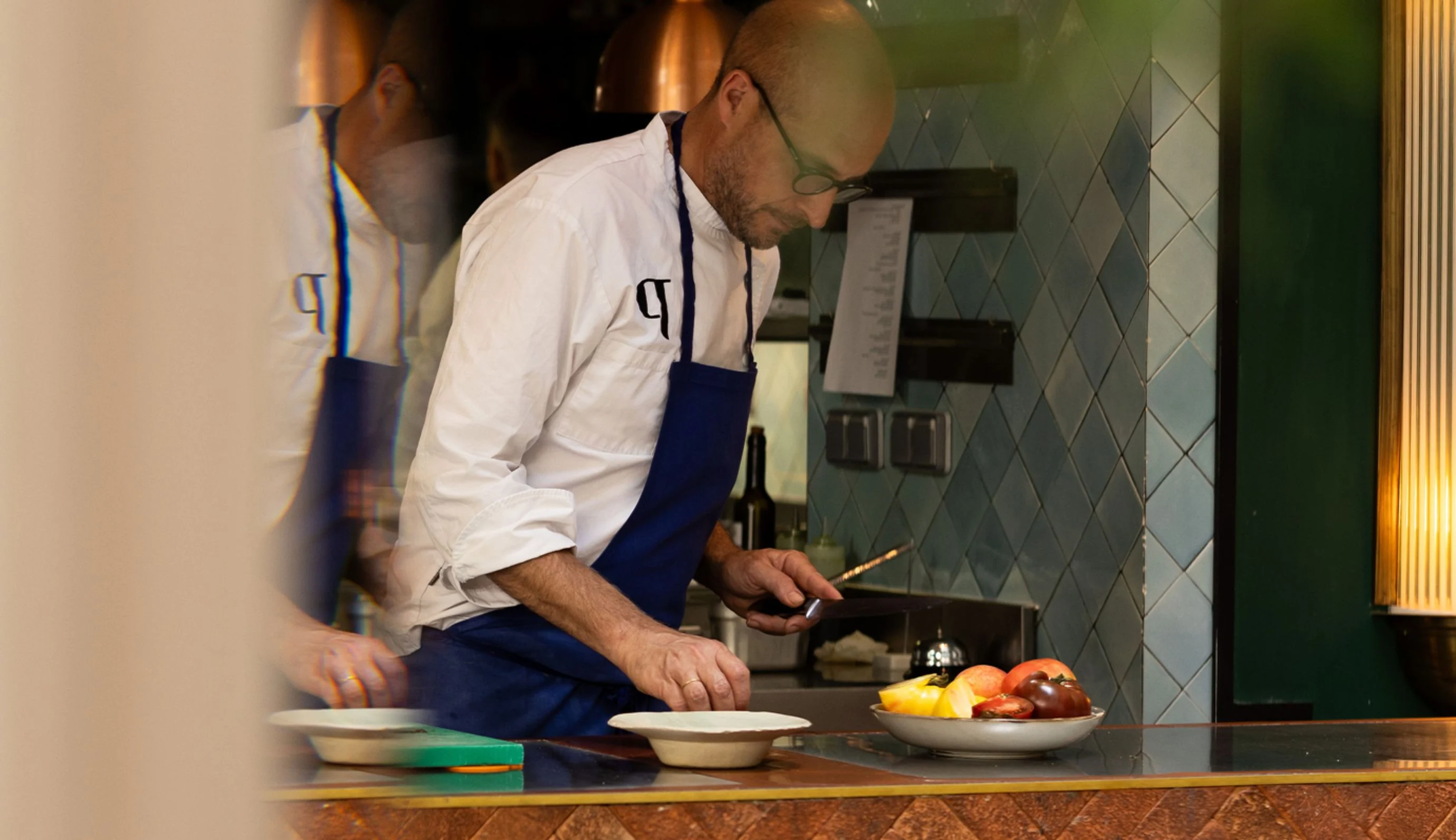 Chef in white shirt and blue apron preparing food behind a counter with fresh tomatoes on a plate.