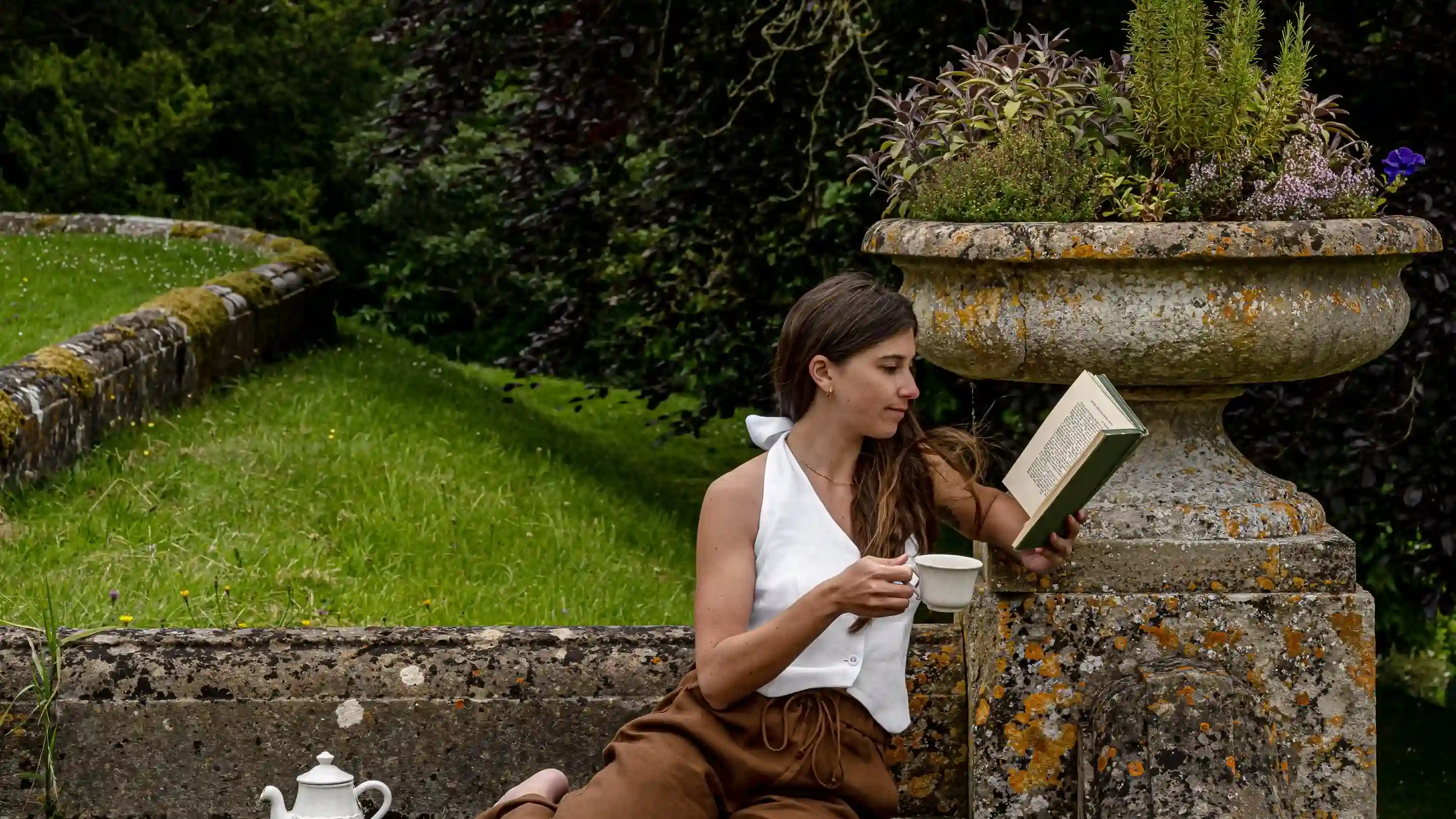 Mujer con cabello largo sentada en una repisa de piedra al aire libre, leyendo un libro y sosteniendo una taza de té cerca de una maceta grande.