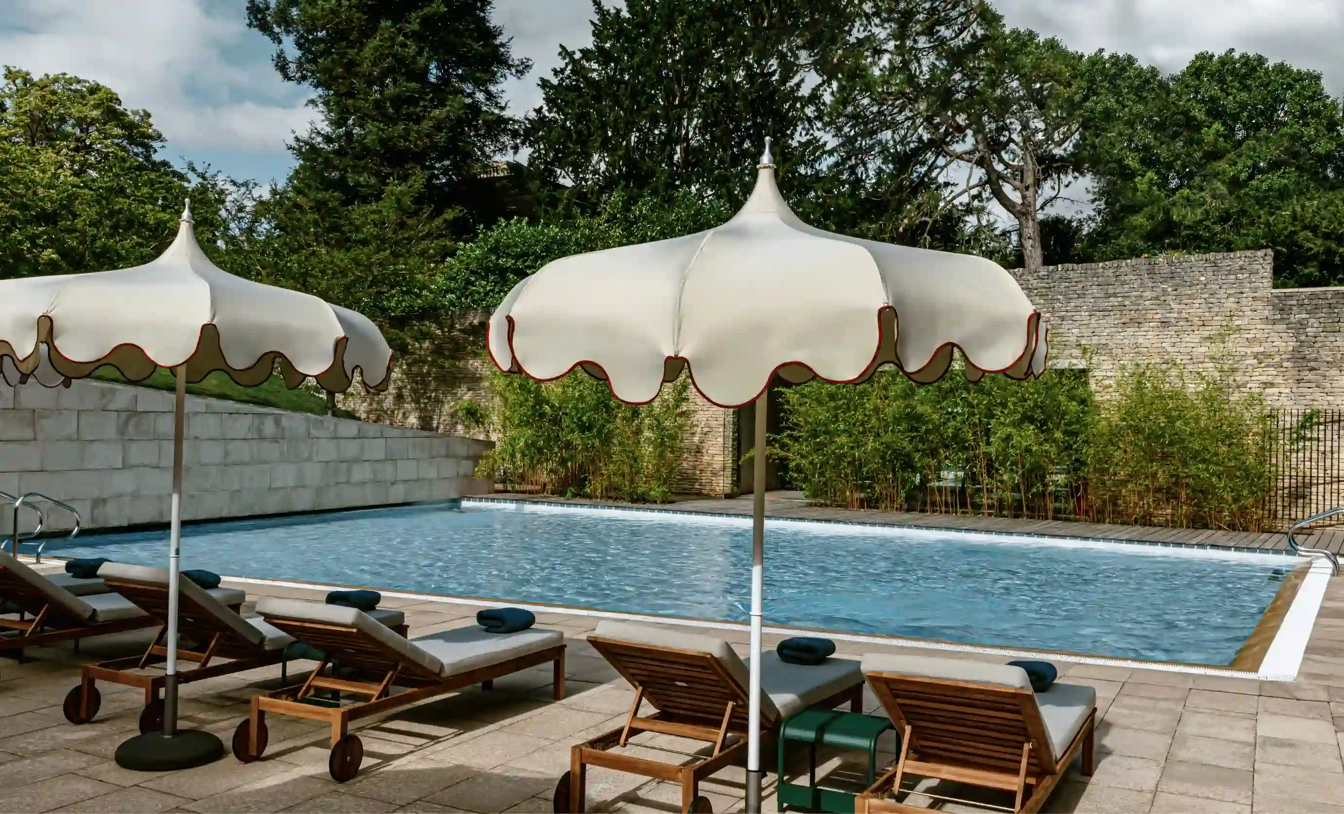 Piscine extérieure avec parasols beiges et chaises longues en bois sur une terrasse carrelée entourée de verdure.
