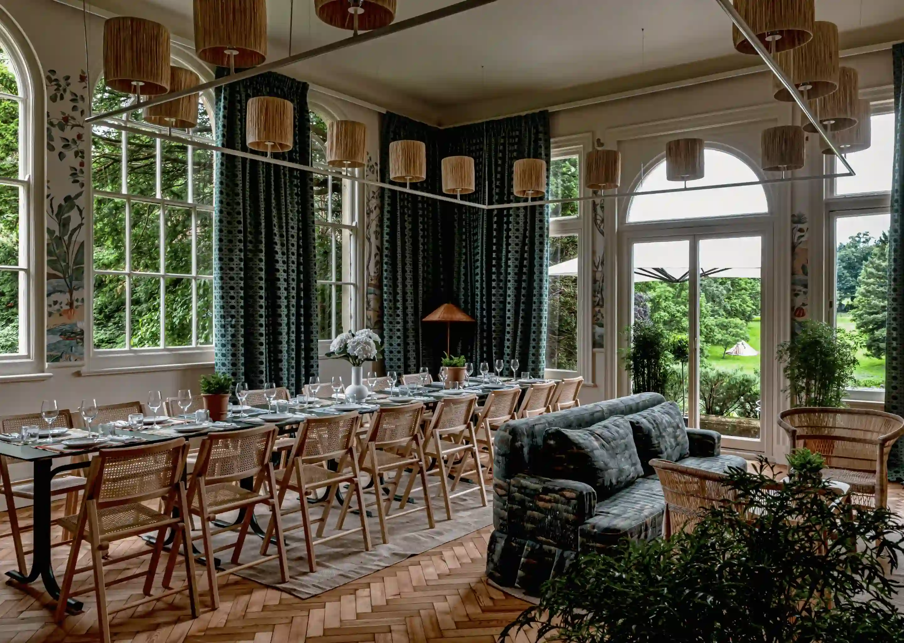 Comedor con mesa larga para invitados, sillas de madera, sofá, plantas y grandes ventanales con vistas al jardín.