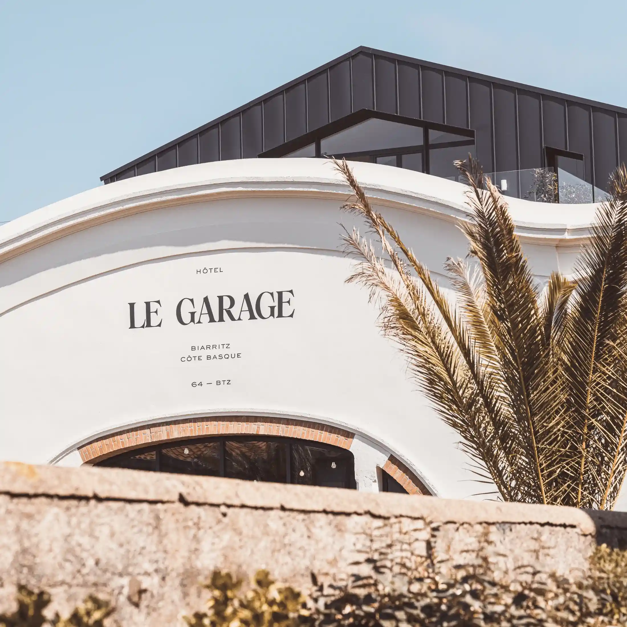 Fachada blanca del edificio con el letrero del hotel «Le Garage» y una palmera alta al lado bajo un cielo despejado.
