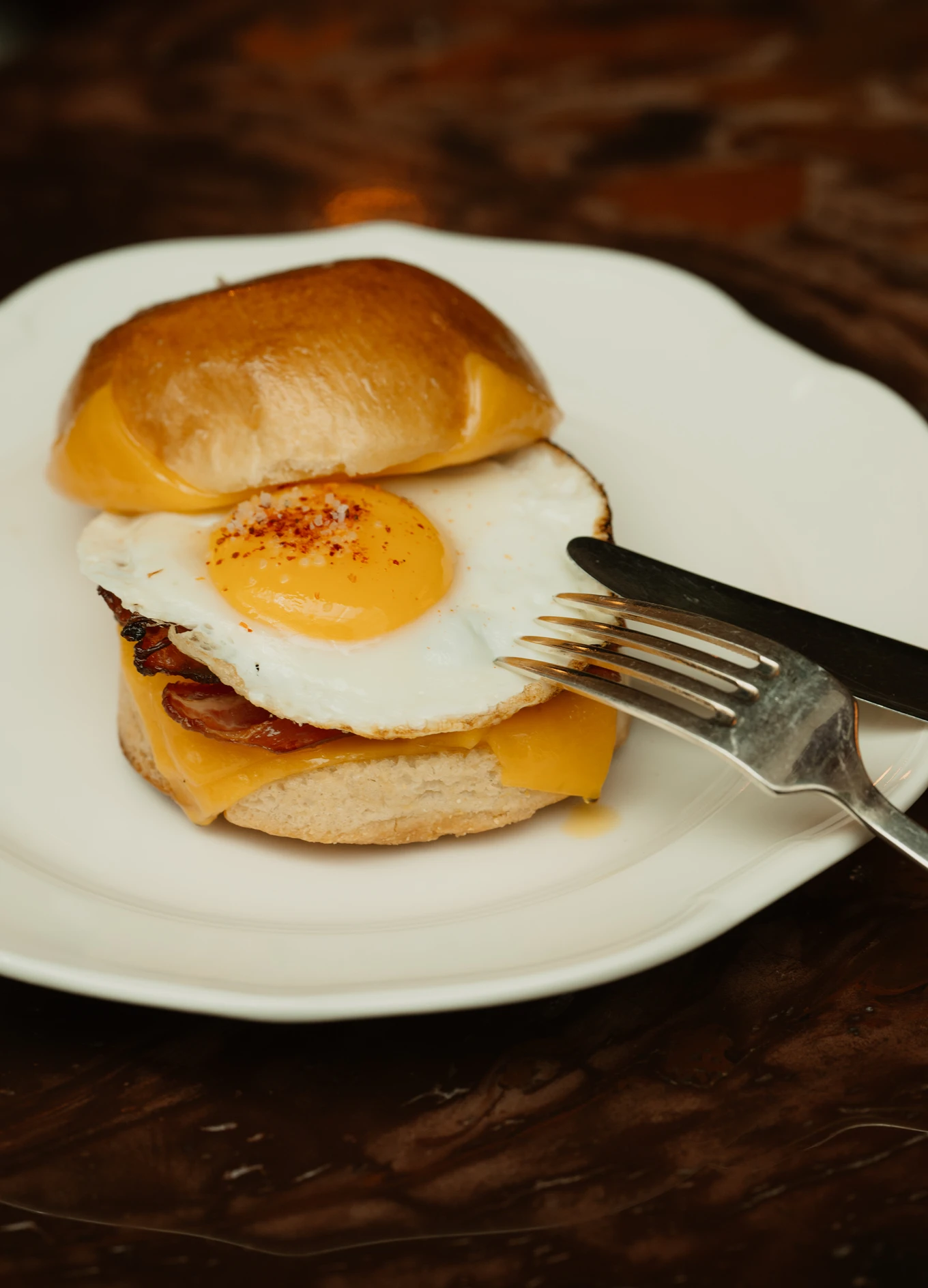 Panino per la colazione con uova fritte, formaggio cheddar e pancetta su un piatto bianco con forchetta e coltello.