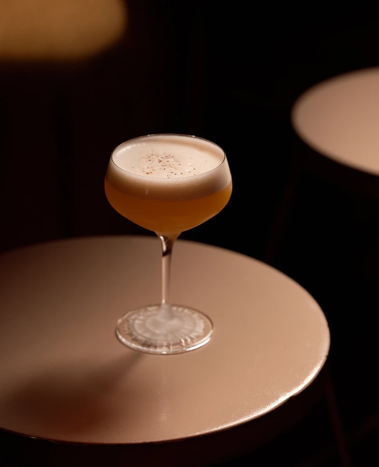 Coquetel com espuma e especiarias polvilhados por cima em um copo cupê sobre uma mesa redonda bege.