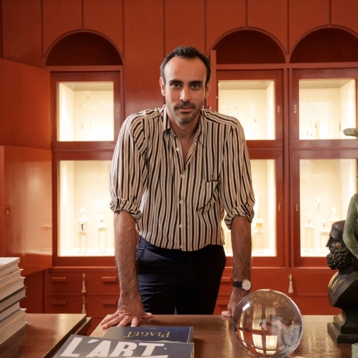 Homem de camisa listrada apoiado em uma mesa com livros de arte e esculturas atrás dele em uma sala de galeria.