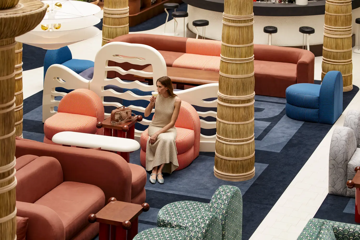 Woman in beige dress sitting on orange striped chair, sipping drink in colorful modern lounge area.
