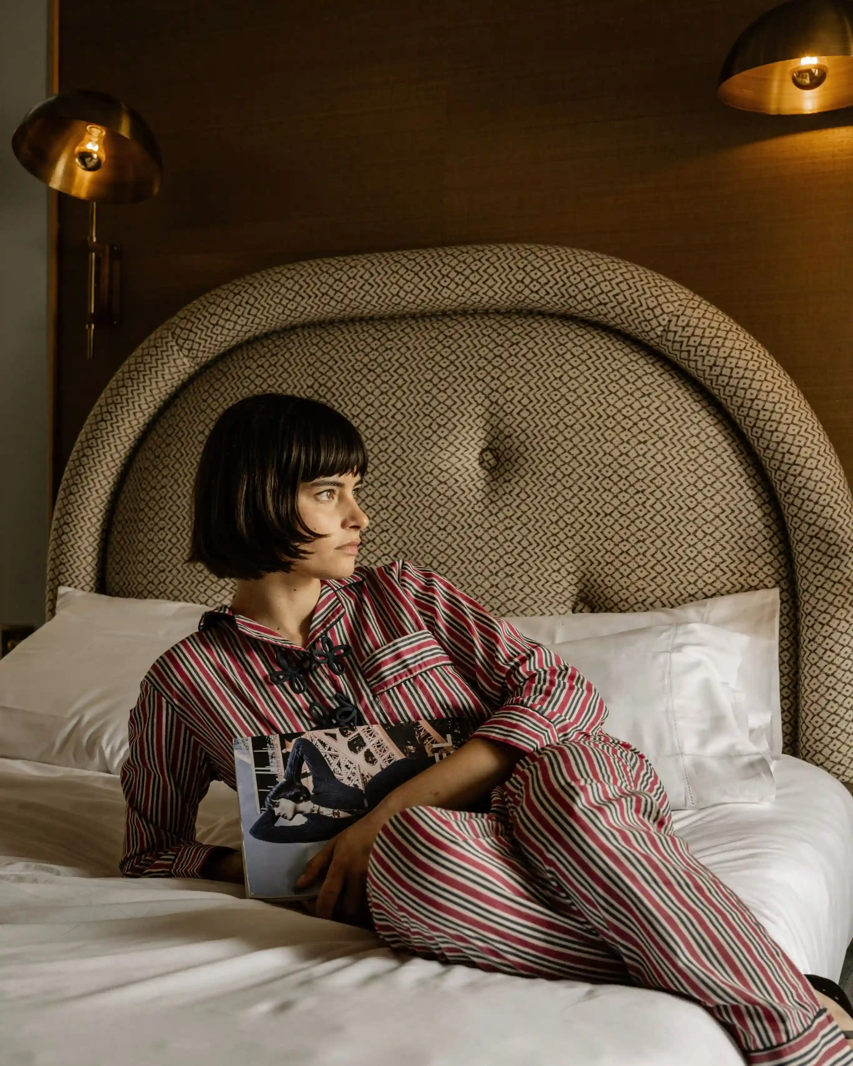 Pessoa de pijama listrado reclinada em uma cama segurando um livro, com cabeceira estampada e lâmpadas quentes.