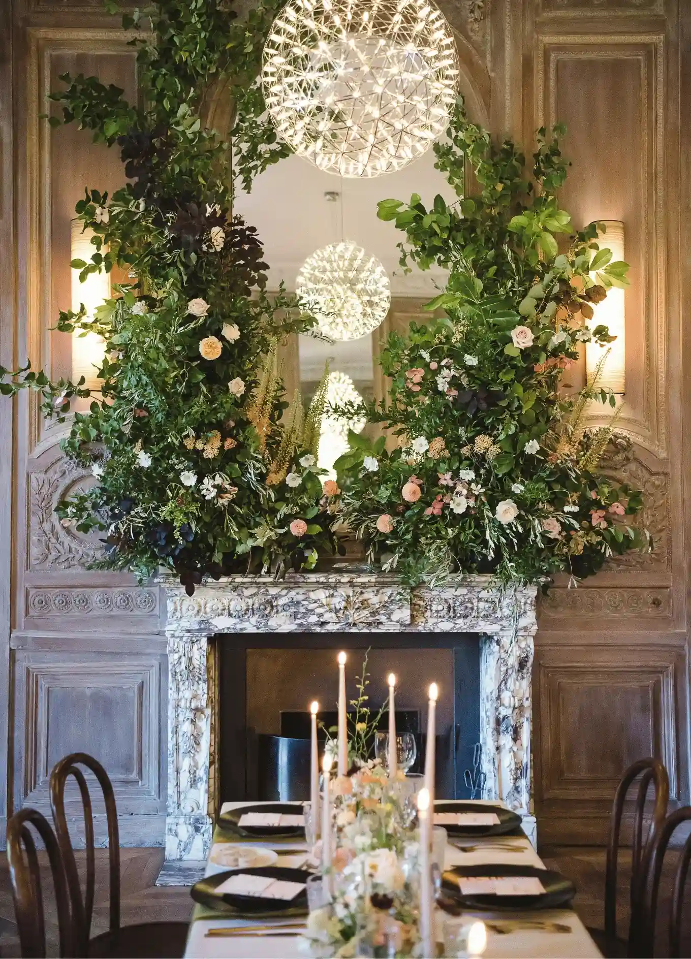 Elegante mesa de comedor con velas encendidas frente a un gran arreglo floral sobre una chimenea de mármol.