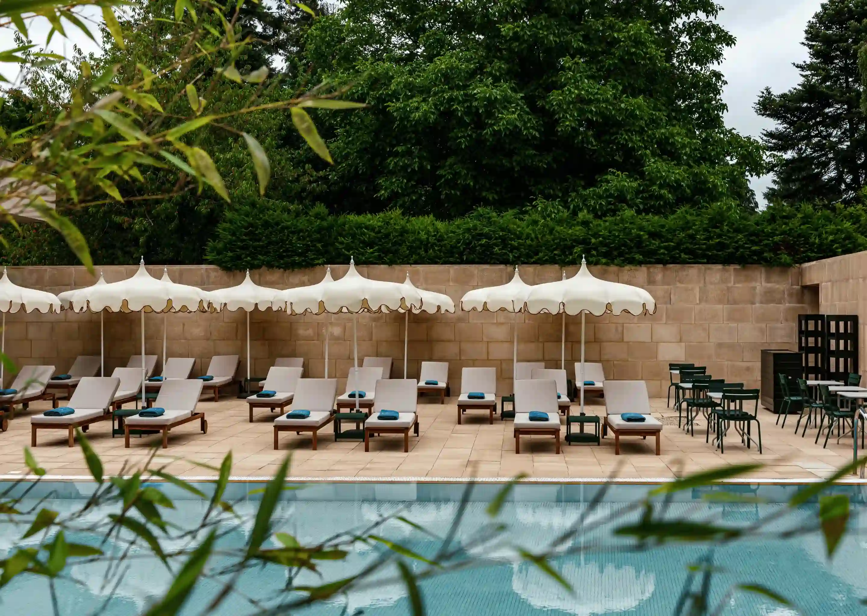 Espace vide au bord de la piscine avec des chaises longues bordées, des parasols blancs et des arbres verts derrière un mur de pierre.