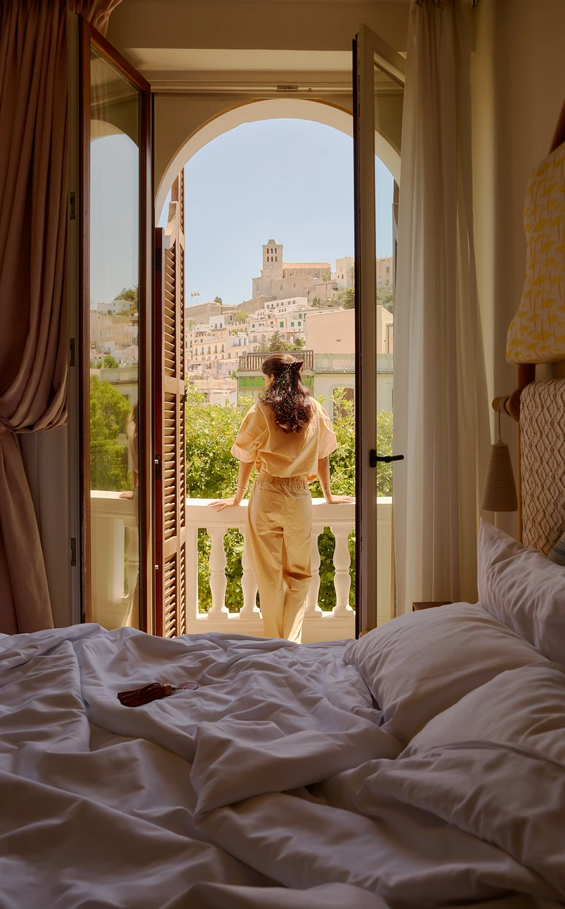 Personne en tenue beige debout sur le balcon regardant les bâtiments à flanc de colline et la tour en plein jour.