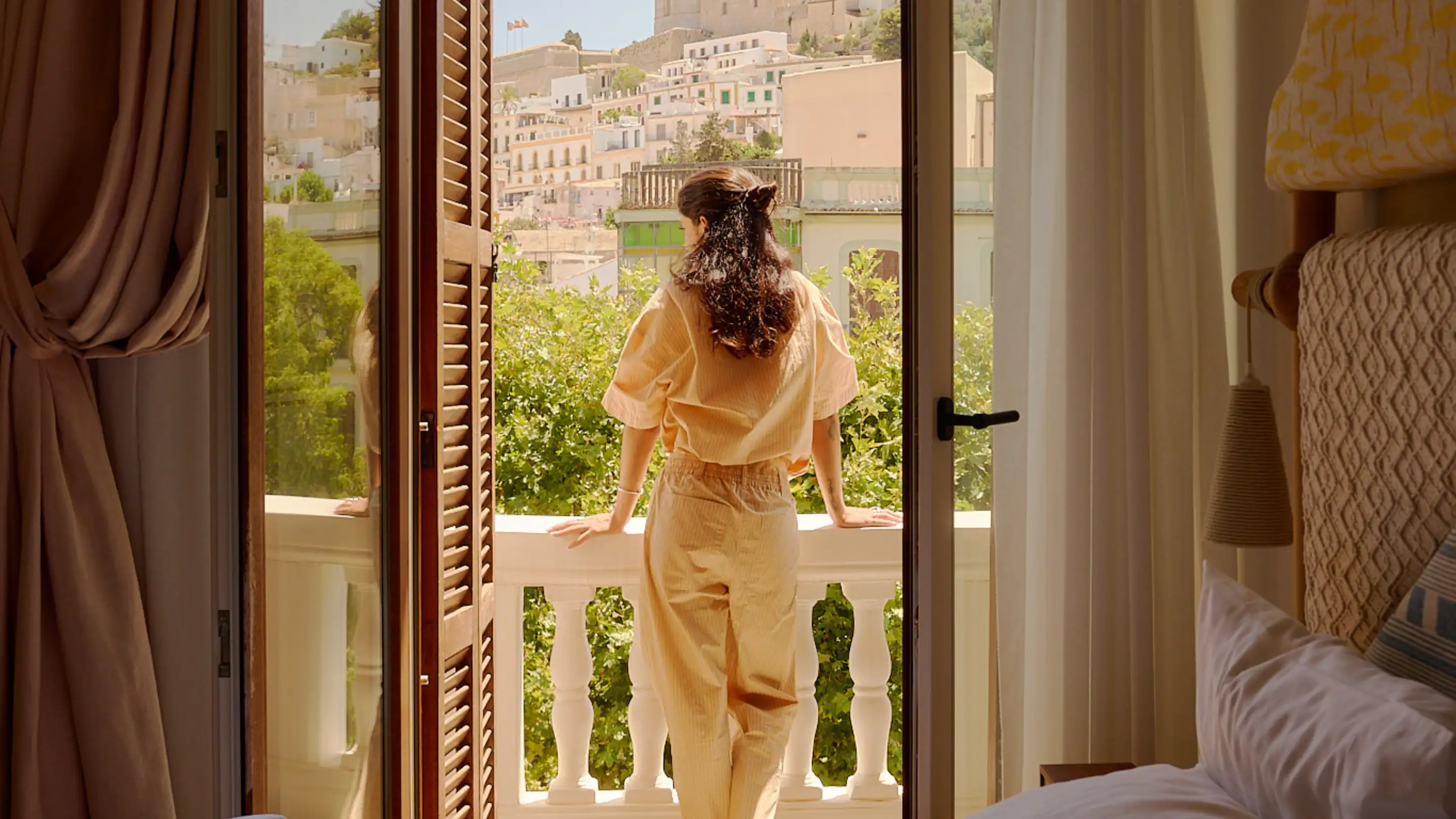 Femme en vêtements beiges debout sur un balcon surplombant des arbres verts et des bâtiments blancs par une journée ensoleillée.