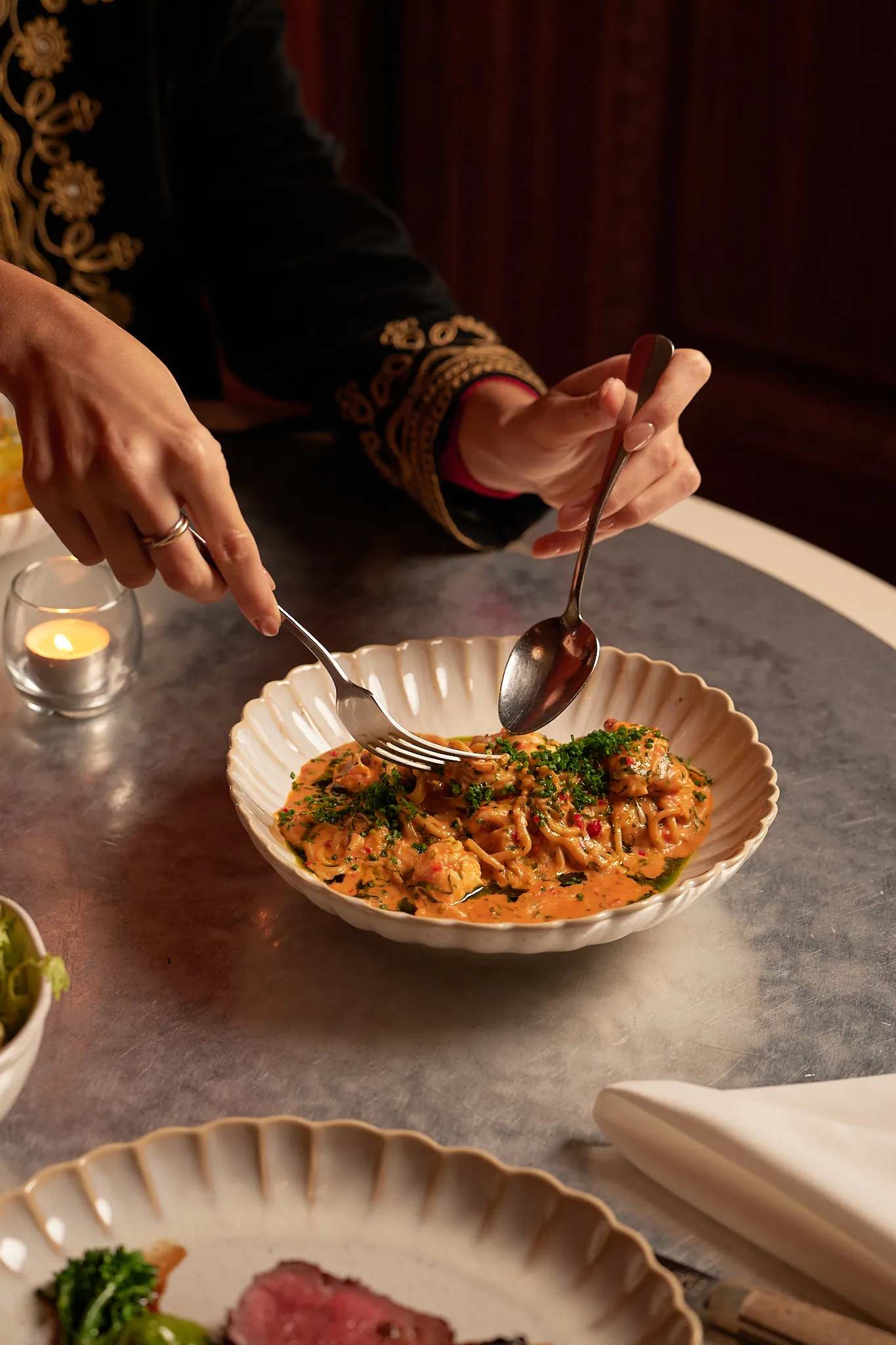 Mani che usano forchetta e cucchiaio per mangiare pasta cremosa condita con erbe aromatiche su un tavolo rotondo con candela.