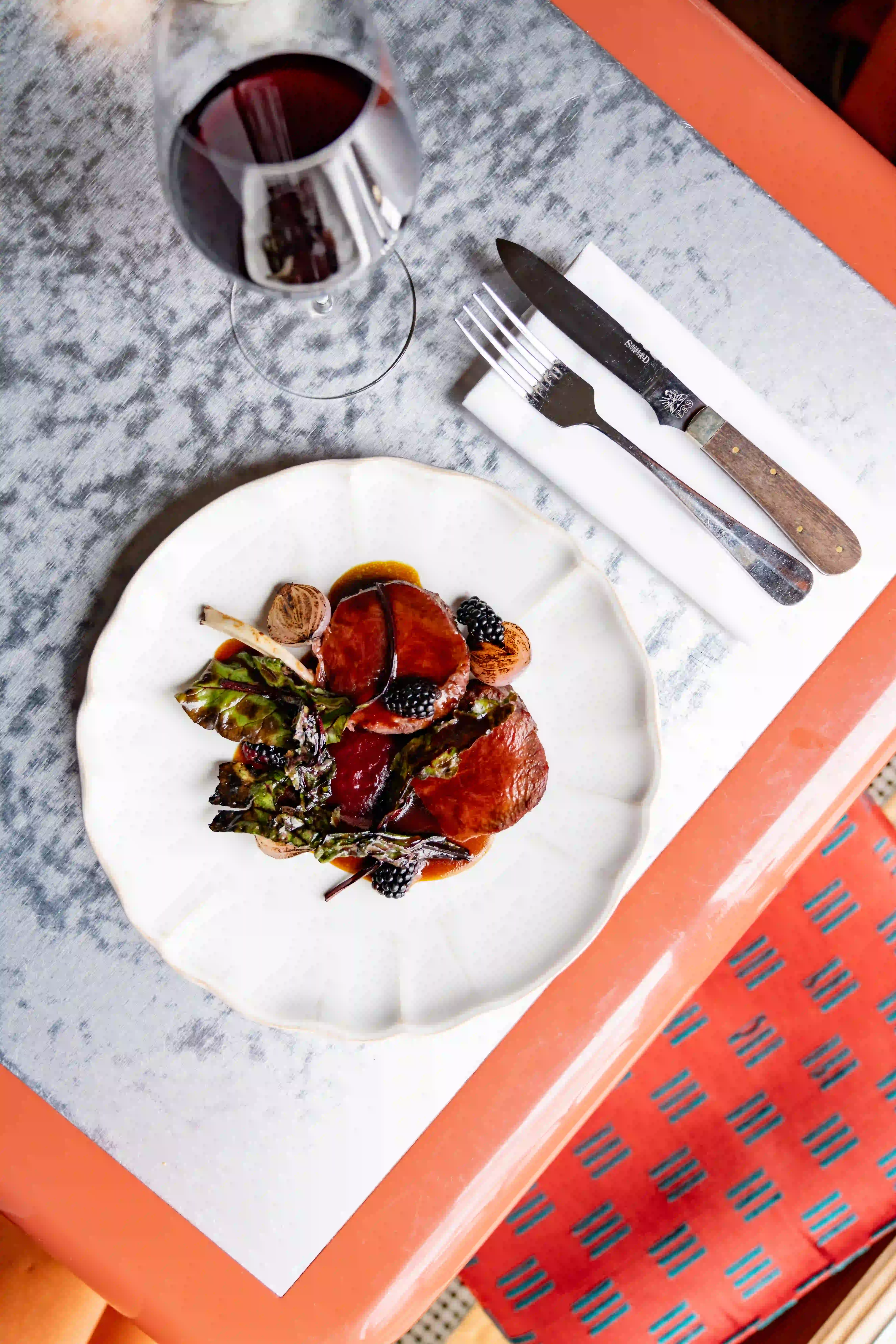 Piatto con carne gourmet, verdure a foglia verde, more, noci e salsa accanto a un bicchiere di vino e posate su un tavolo.