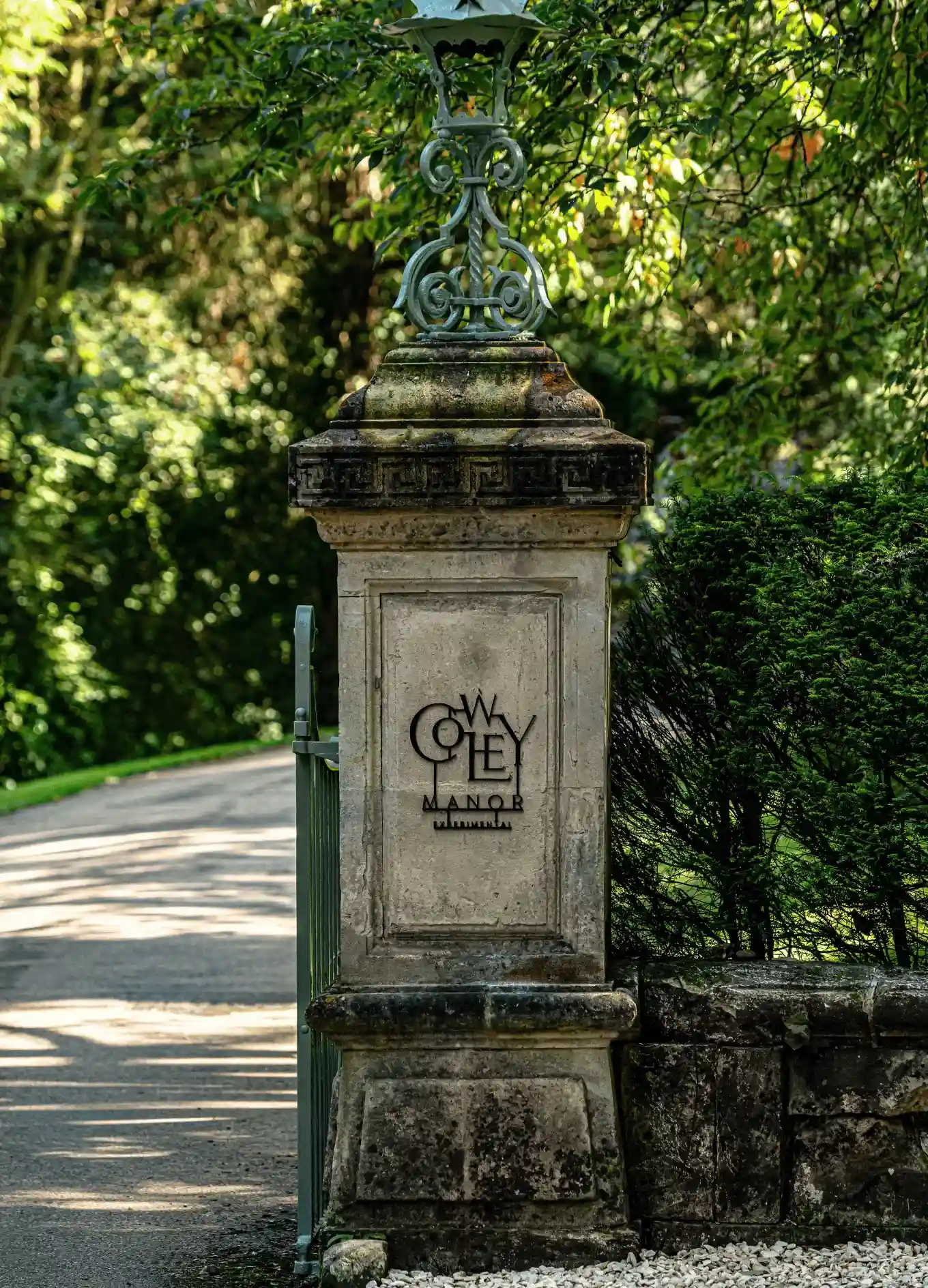 Pilastro in pietra con una lampada in metallo decorato e l'insegna di Cowley Manor vicino a un cancello verde e fogliame lussureggiante.