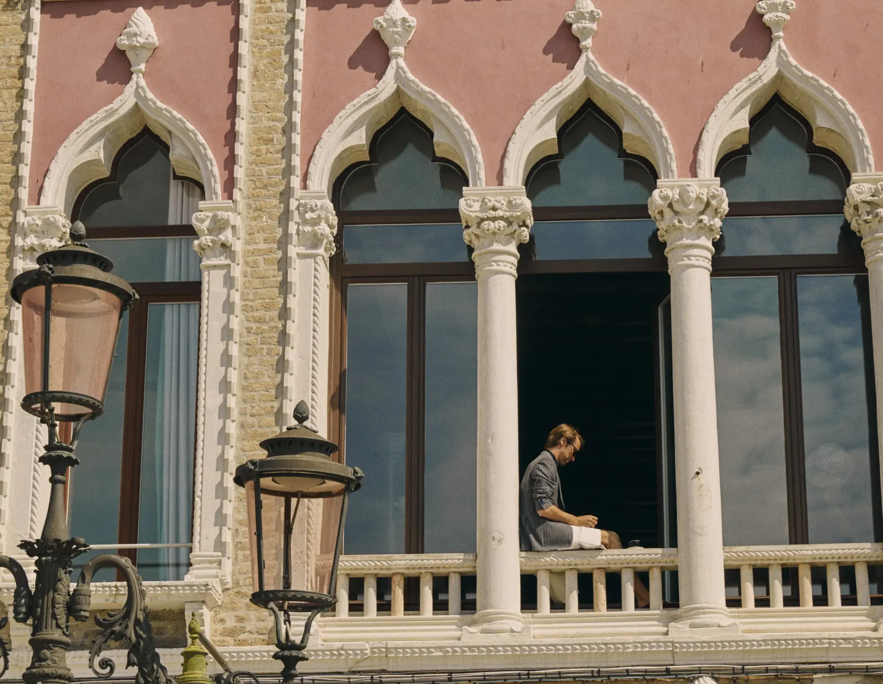 Person sitting on ornate balcony of historic building with arched windows and decorative columns.
