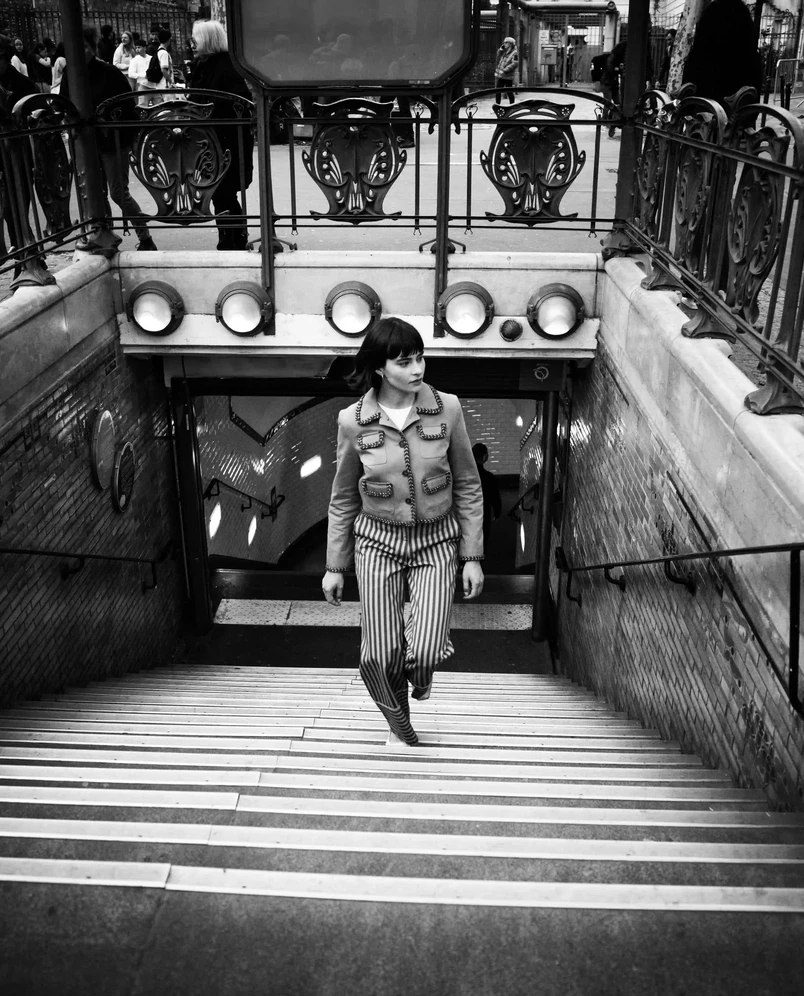 Femme en pantalon rayé et veste montant les escaliers depuis une entrée de métro sur une photo en noir et blanc.
