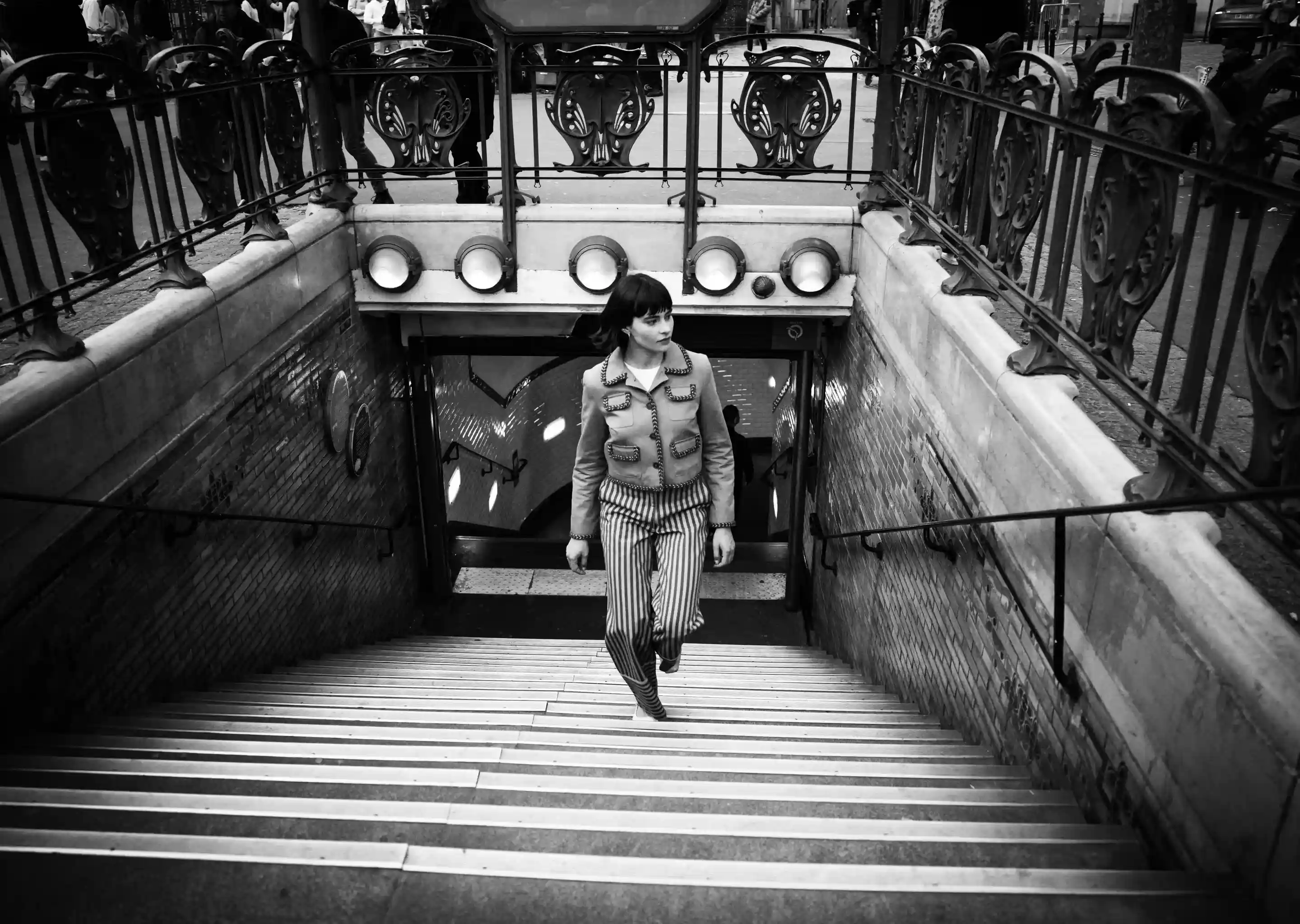 Femme en pantalon rayé et veste montant les marches d'une entrée de métro avec des balustrades ornées.