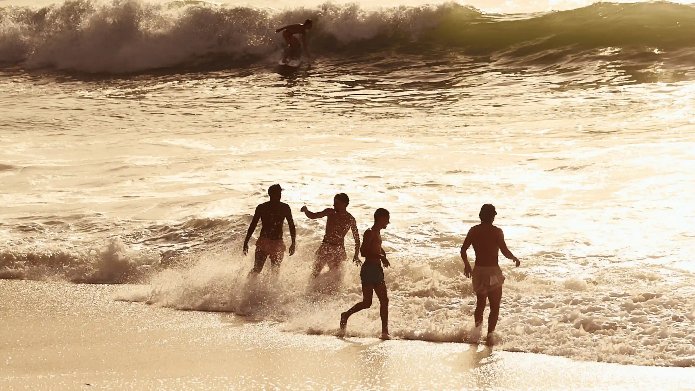 Quattro persone giocano tra le onde poco profonde dell'oceano al tramonto mentre il surfista cavalca un'onda sullo sfondo.