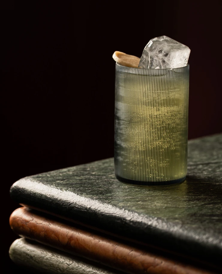 Un bicchiere strutturato con una bevanda verde chiaro, un grosso cubetto di ghiaccio e una scorza di agrumi su una superficie di marmo scuro.