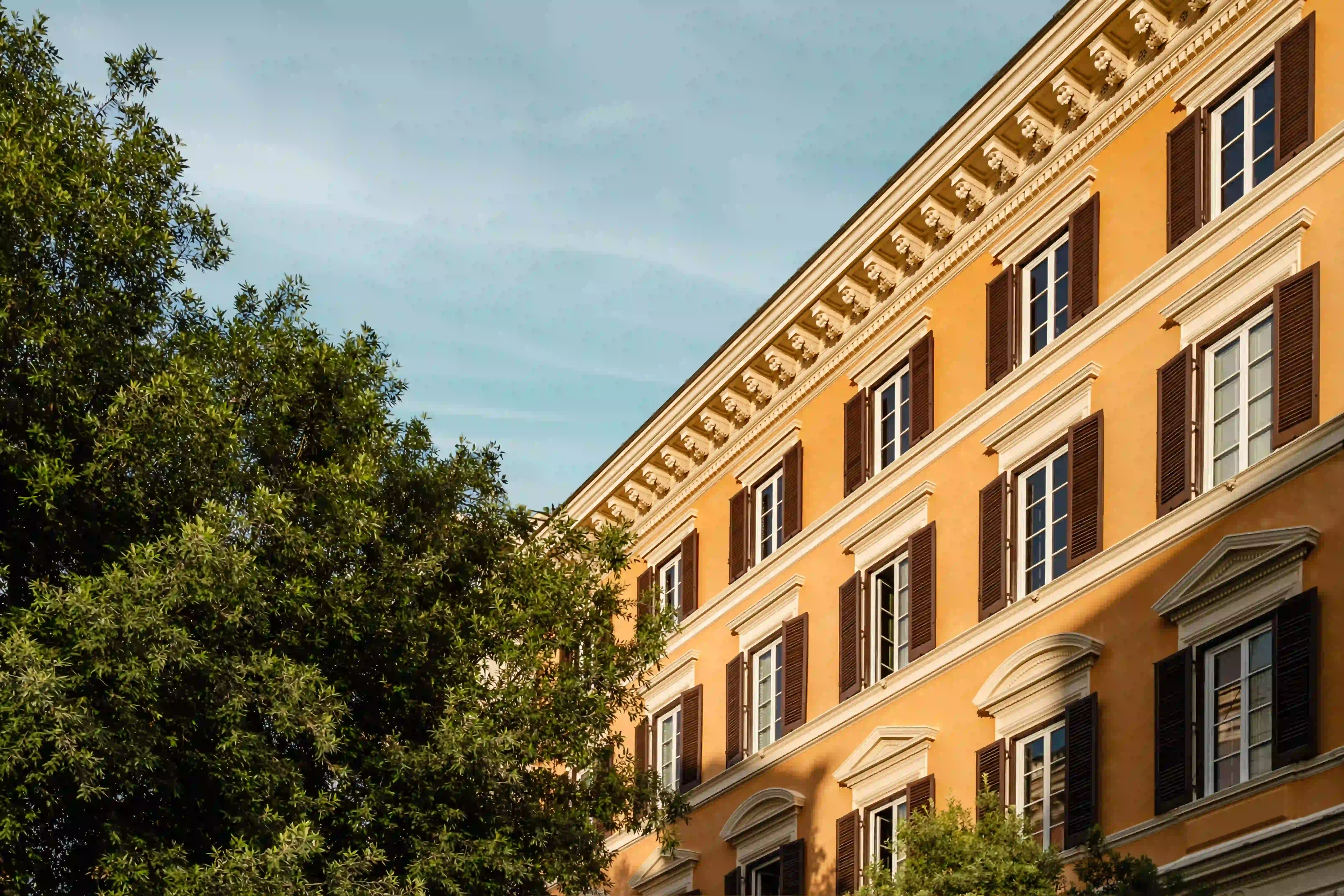 Edificio classico arancione con infissi bianchi e persiane scure accanto ad alberi verdi.