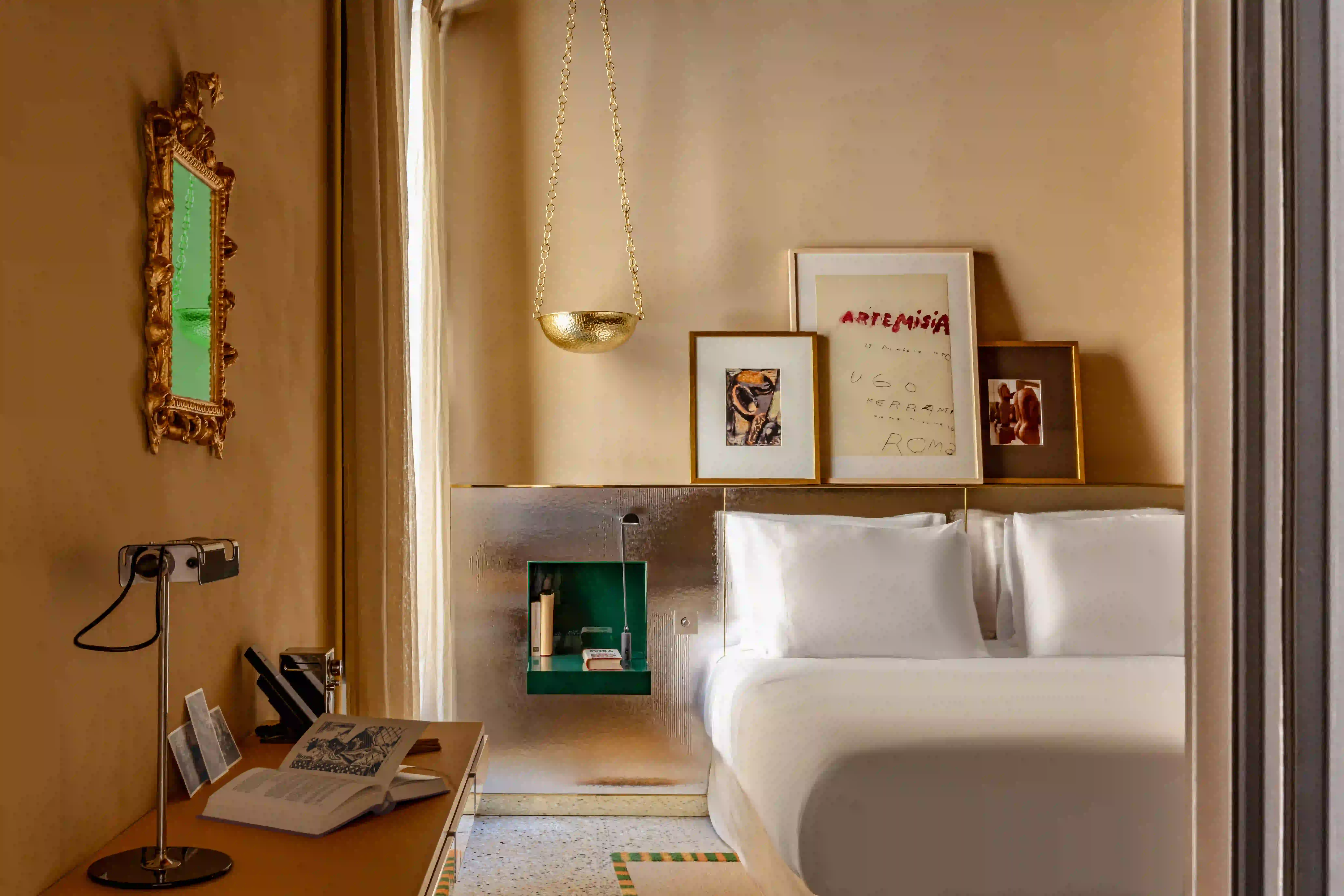 Moderna camera da letto beige con letto bianco, lampada dorata a sospensione, arte murale e scrivania con libro aperto e specchio.