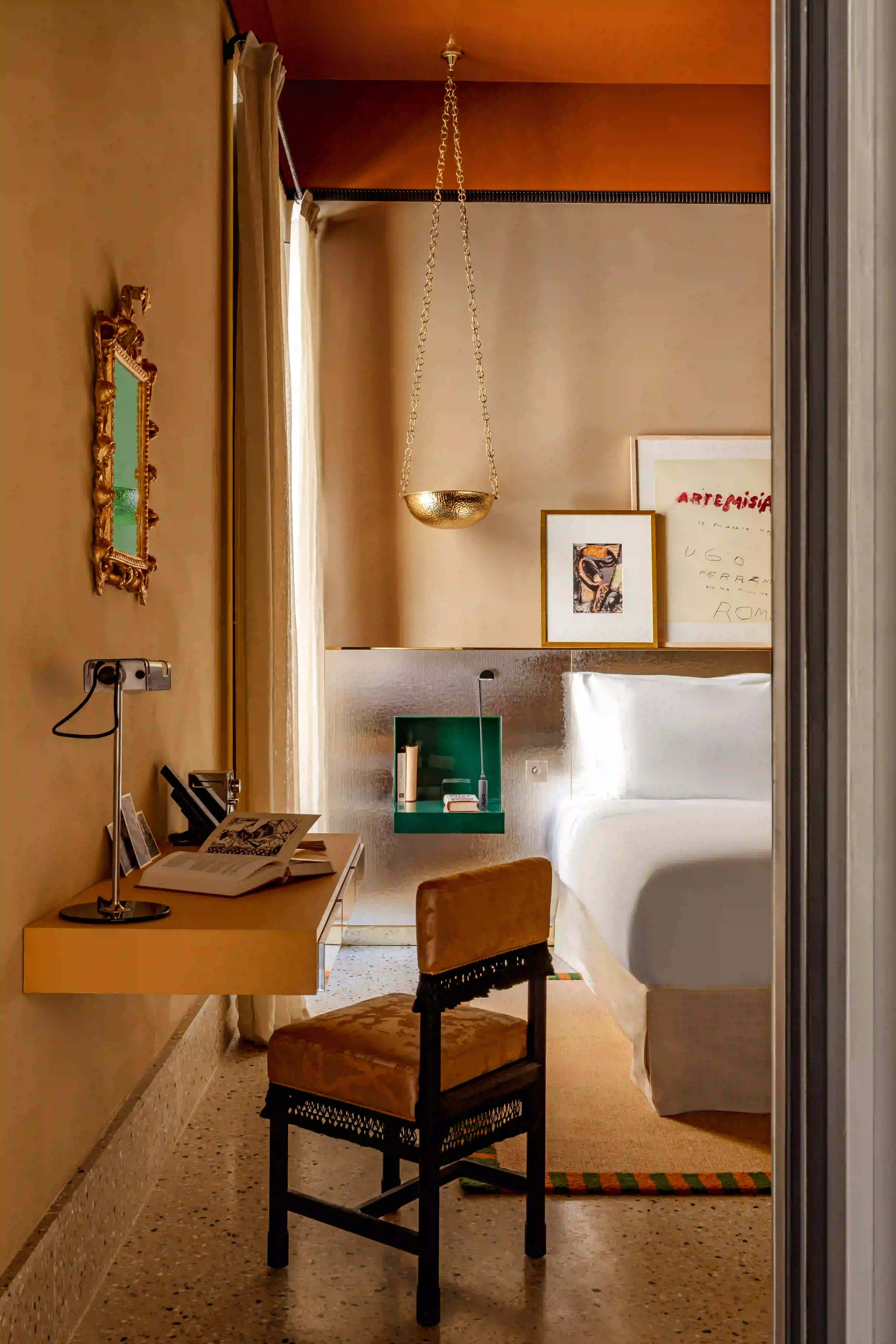 Camera dai toni caldi con scrivania, libro aperto, lampada dorata a sospensione, sedia, letto e opere d'arte incorniciate.