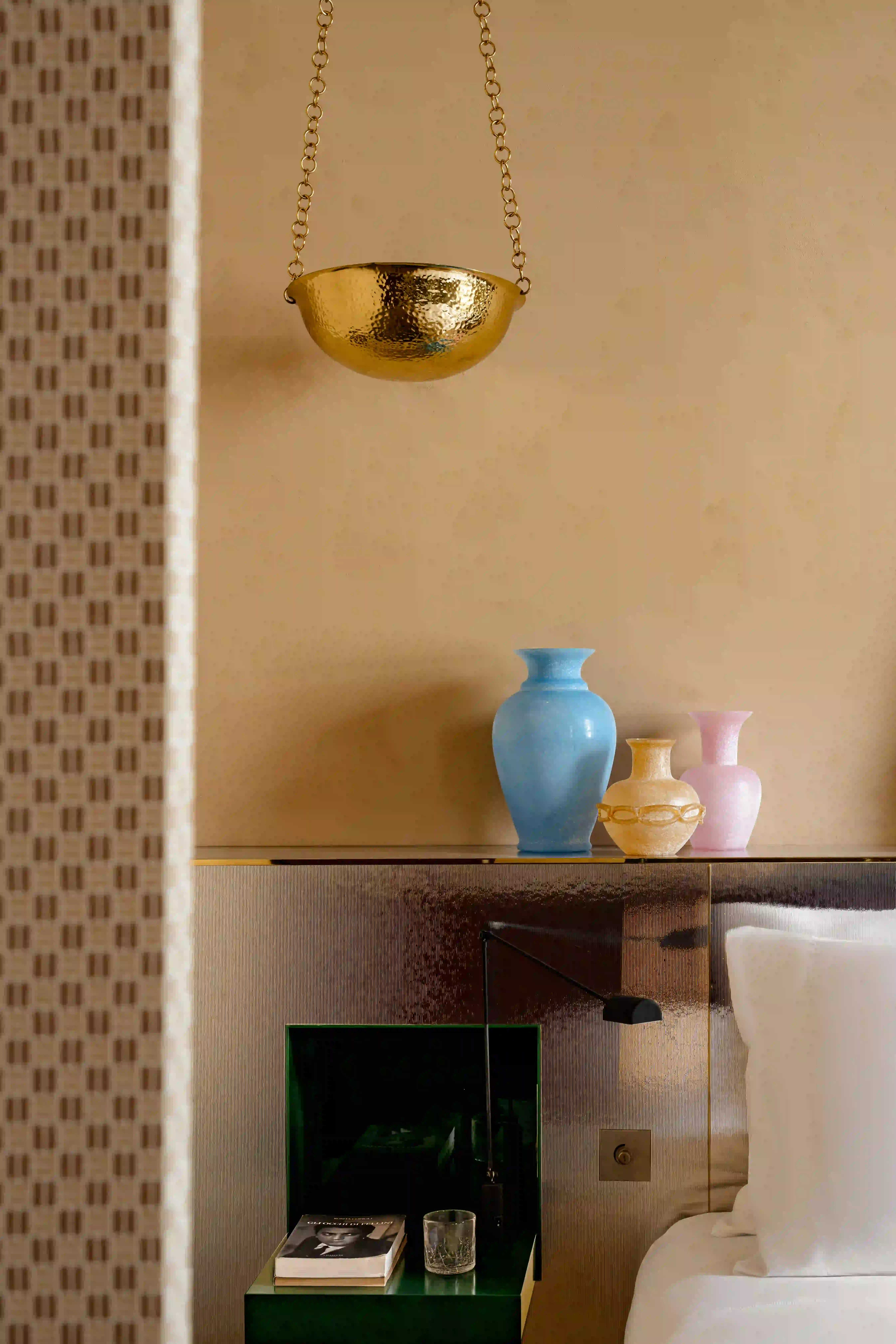 Tre vasi color pastello su una mensola beige sopra un comodino verde con un libro e un bicchiere.