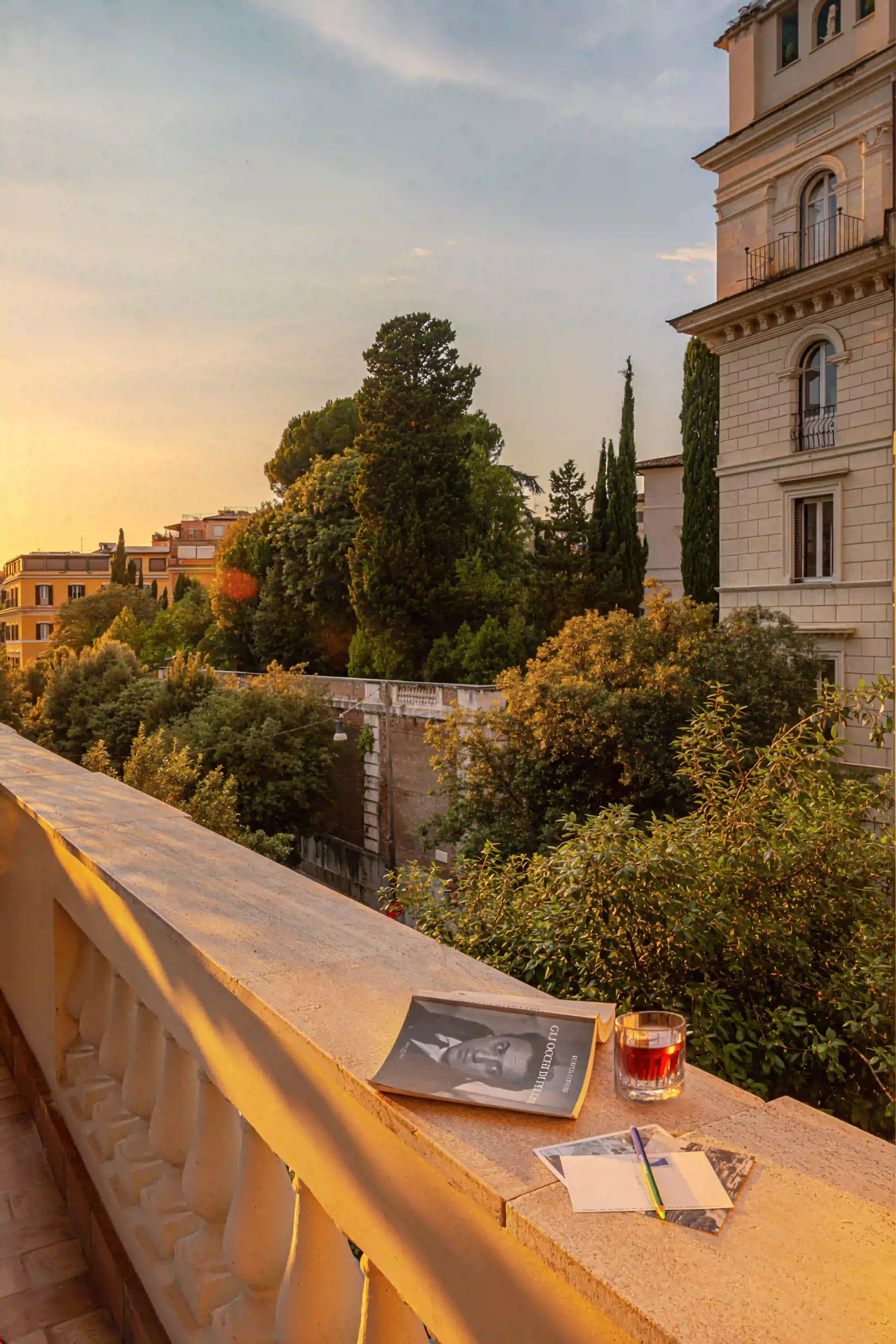 Balcone illuminato dal sole con libro, cartoline, una matita, un bicchiere di bevanda, con vista sul verde degli alberi e degli edifici al tramonto.