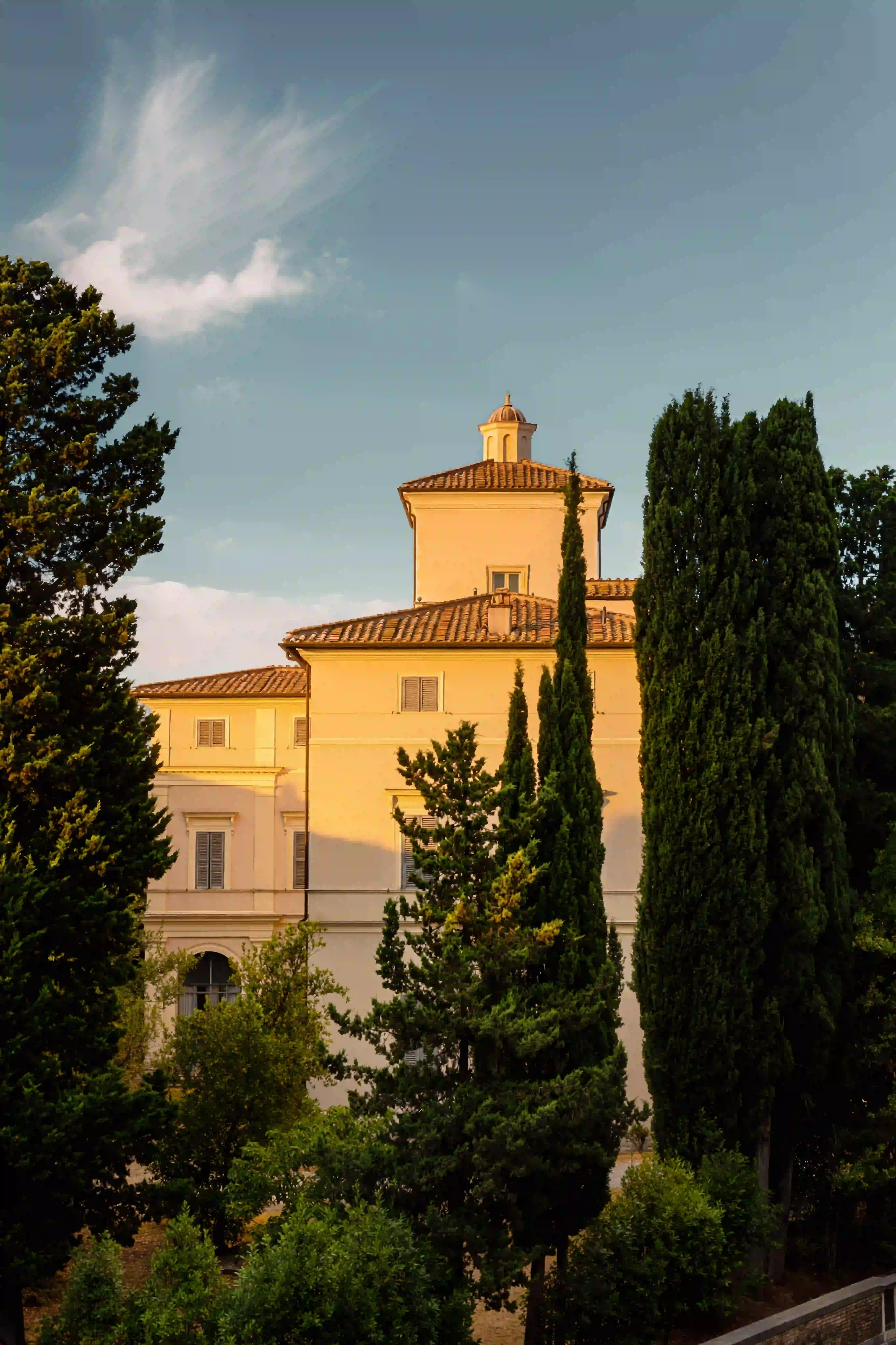 Villa storica con tetto di tegole dietro alti alberi verdi sotto un cielo blu con nuvole sottili.