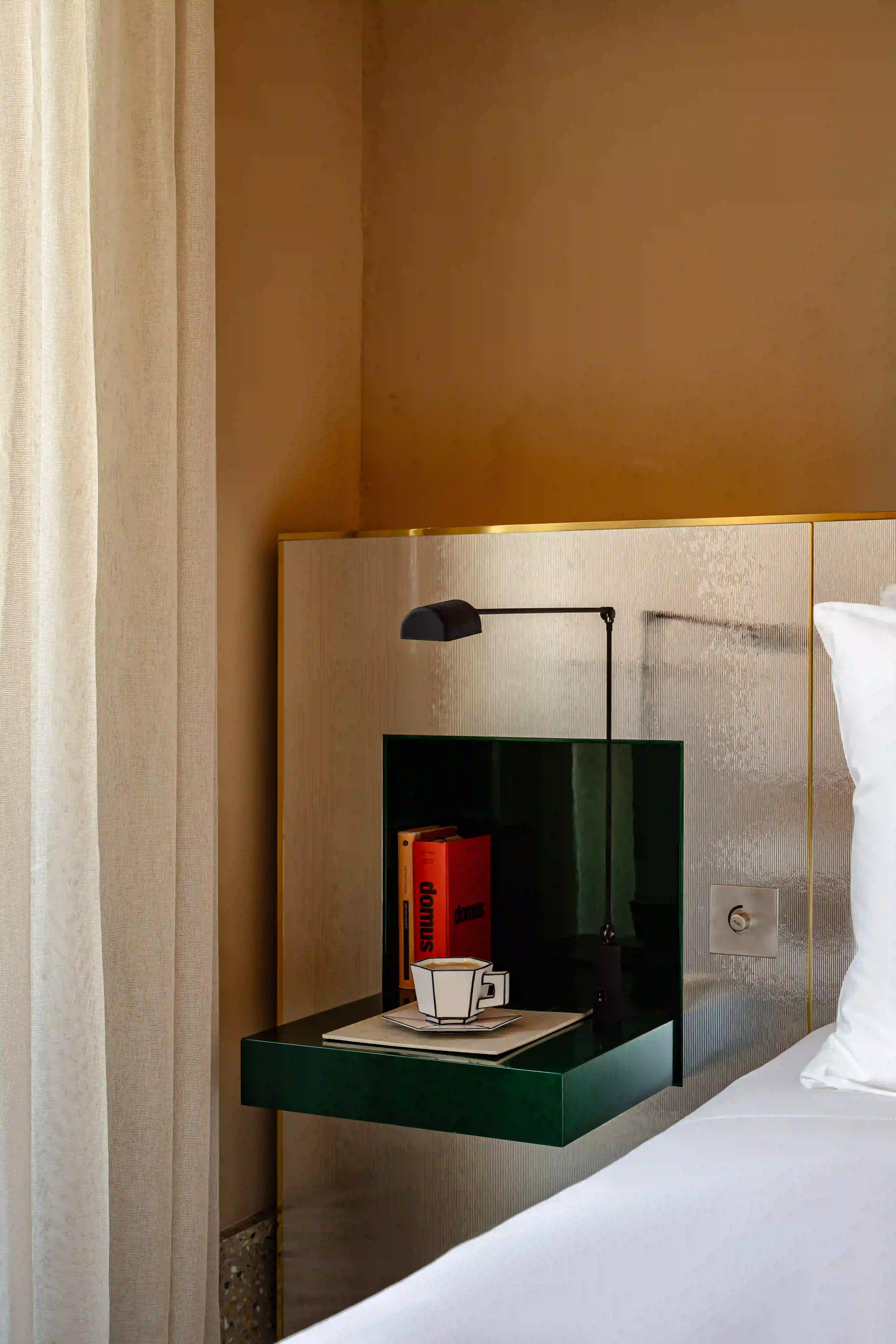 Mesita de noche flotante con libros, una moderna lámpara negra y una taza sobre un platillo cerca de la cama blanca y la cortina.