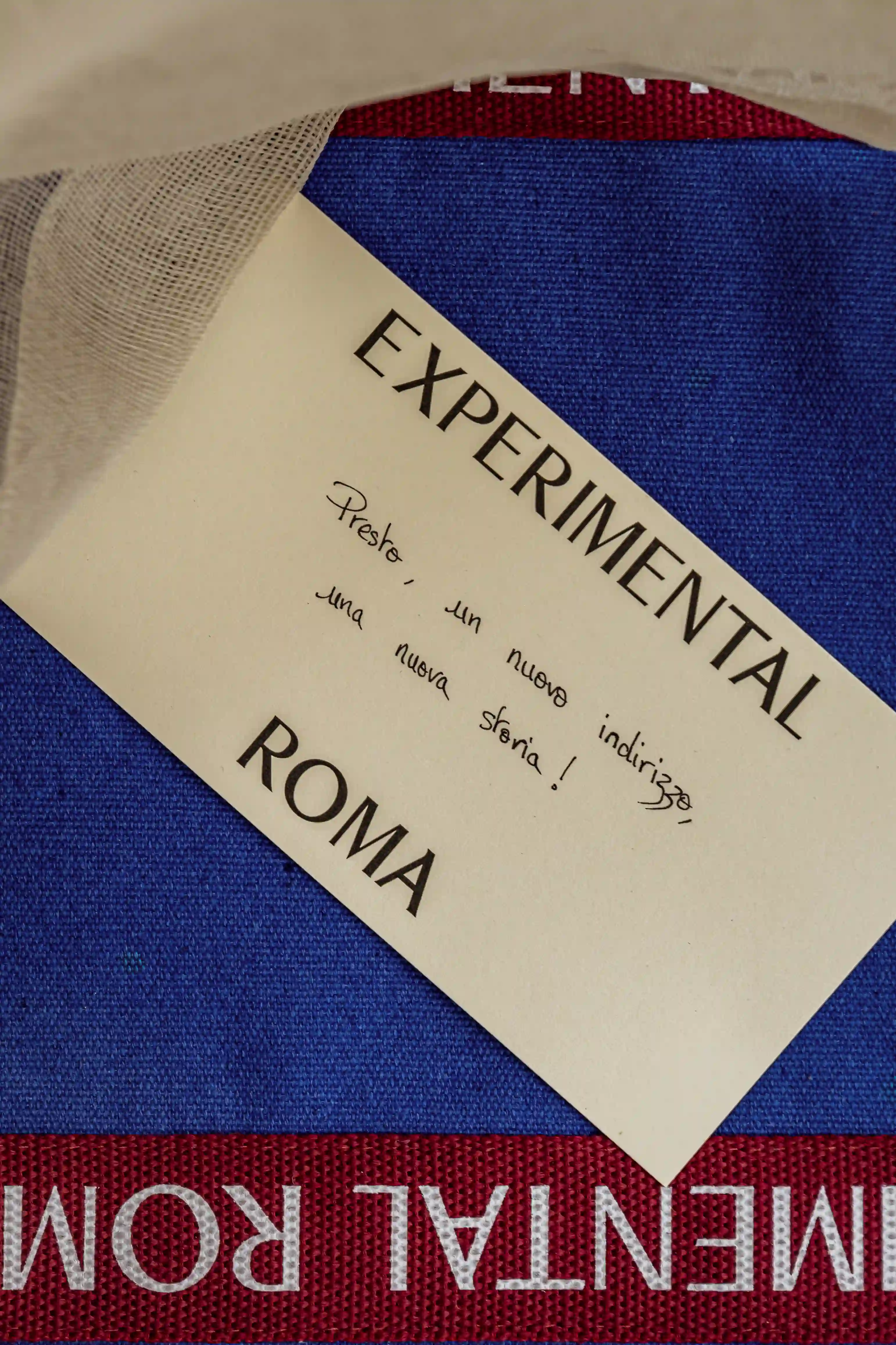 Tarjeta con el texto «EXPERIMENTAL ROMA» sobre tela tejida azul y roja parcialmente cubierta por tela blanca transparente.